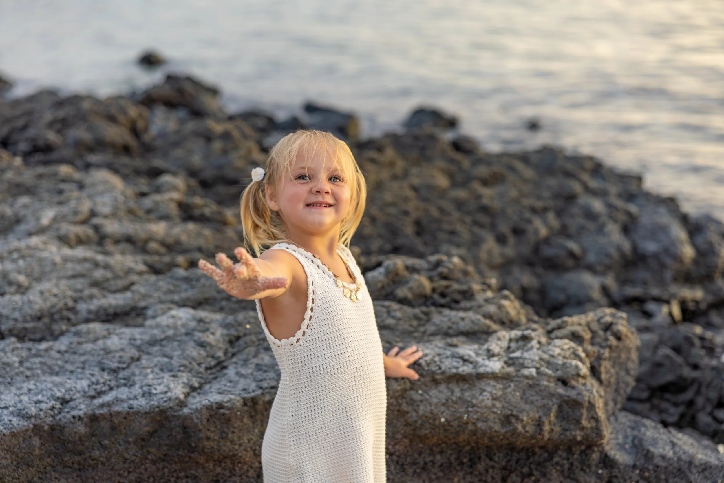 A young girl with blonde hair in pigtails stands on rocks near a body of water, extending her hand towards the camera.