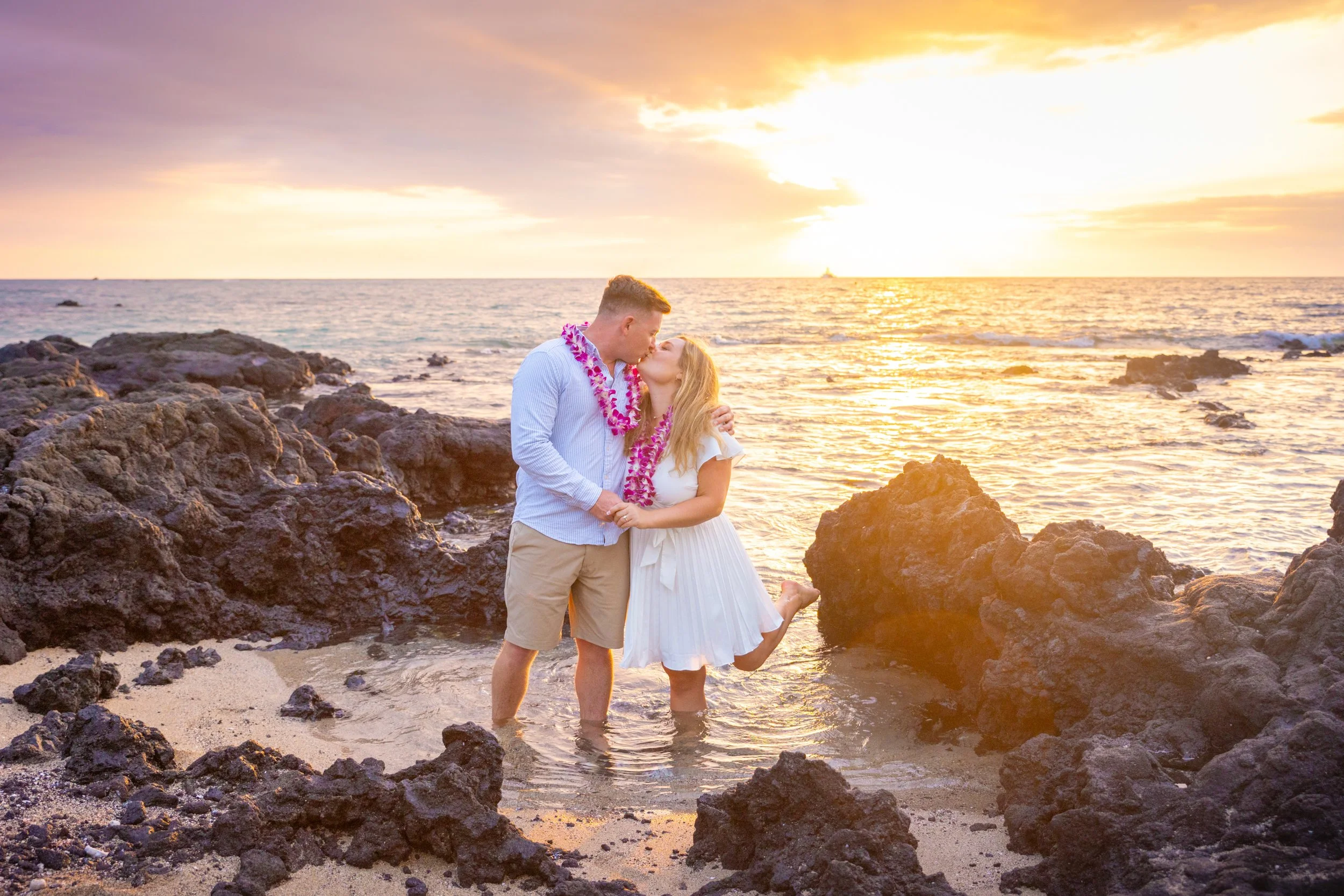 A couple dressed in casual beach attire, holding hands and kissing in the ocean water during sunset, surrounded by rocks on a sandy beach.