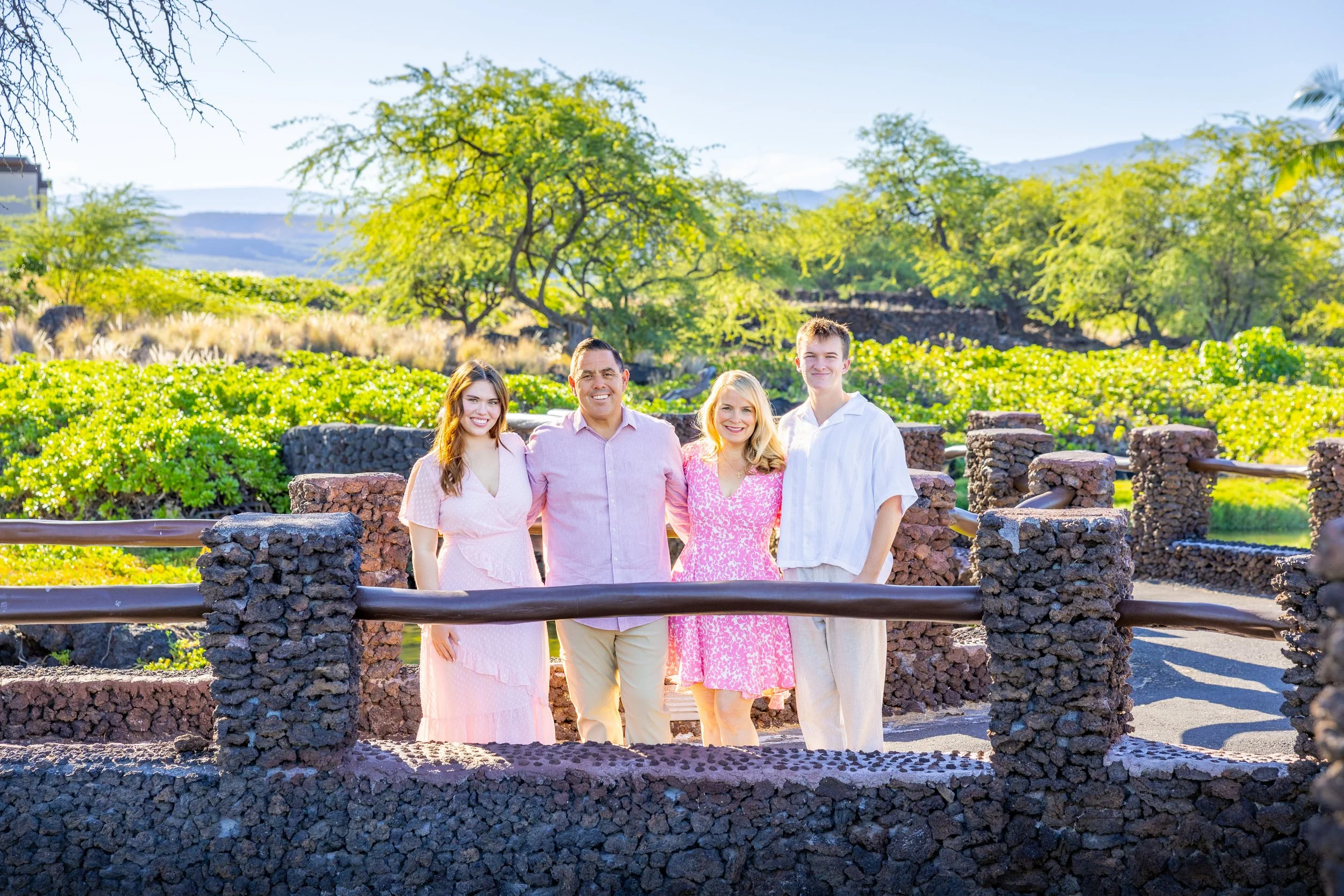 Four people standing on a stone bridge outdoors with greenery and trees in the background, smiling at the camera.