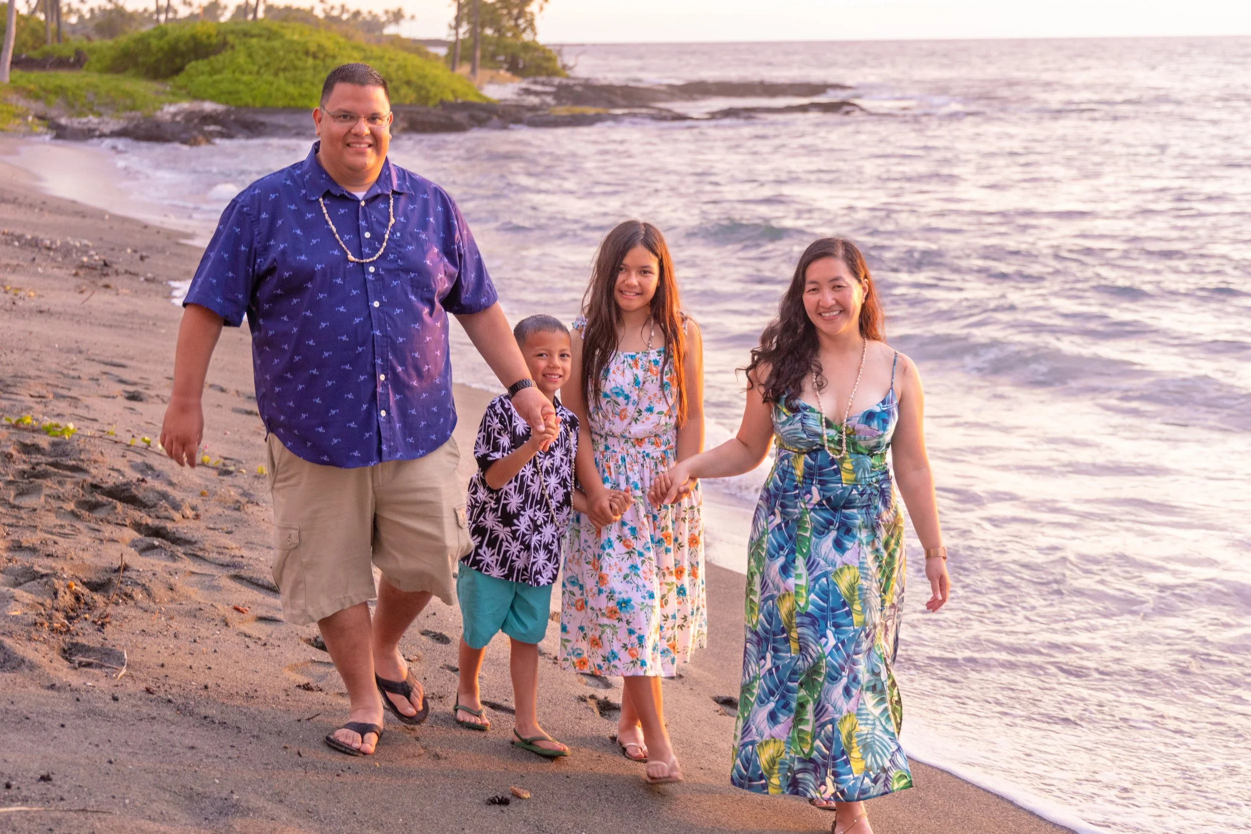 A family of five walking on a beach during sunset, holding hands, dressed in casual tropical clothing, with water and greenery in the background.