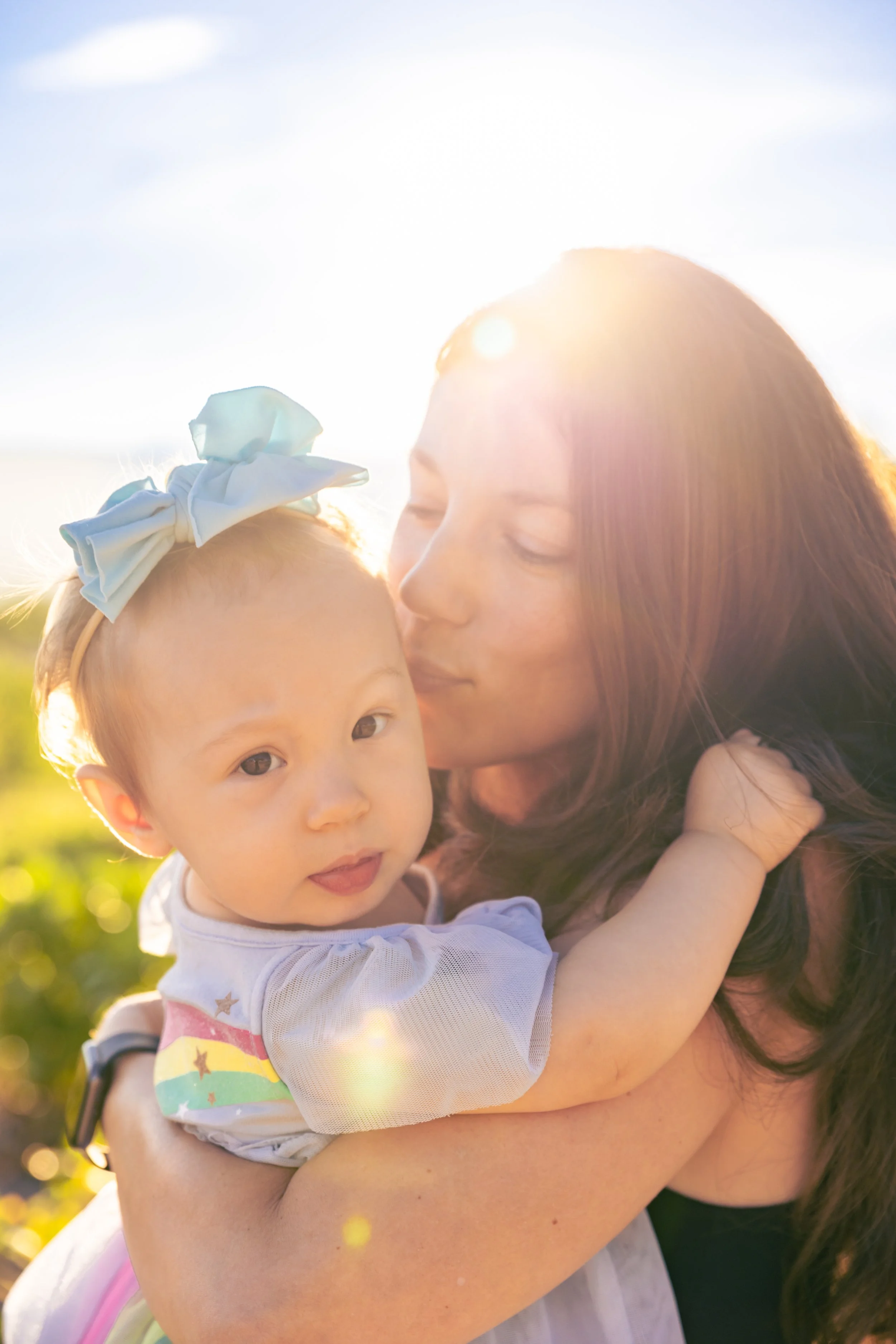 A woman holding a young girl with a rainbow rainbow design on her sleeve, outdoors under bright sunlight.