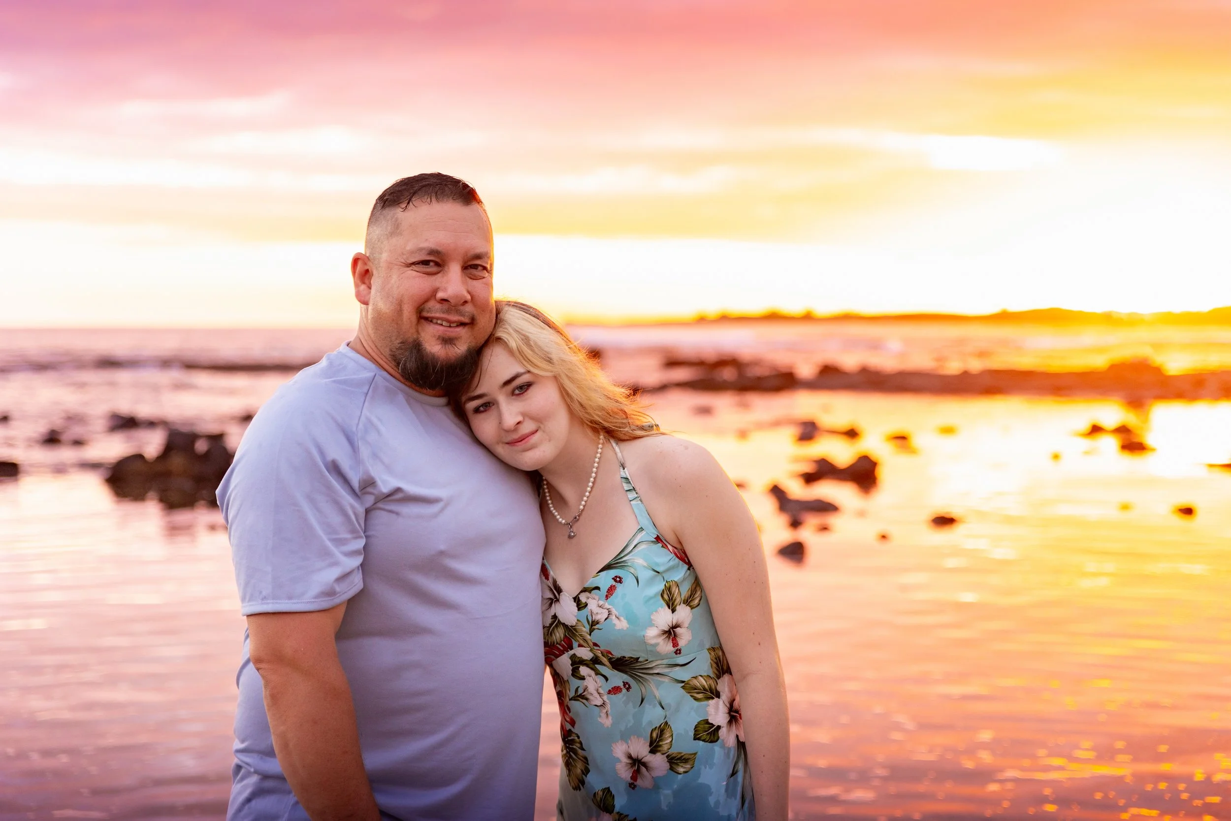 A man and a woman embrace on a beach at sunset, with the ocean and rocks in the background.