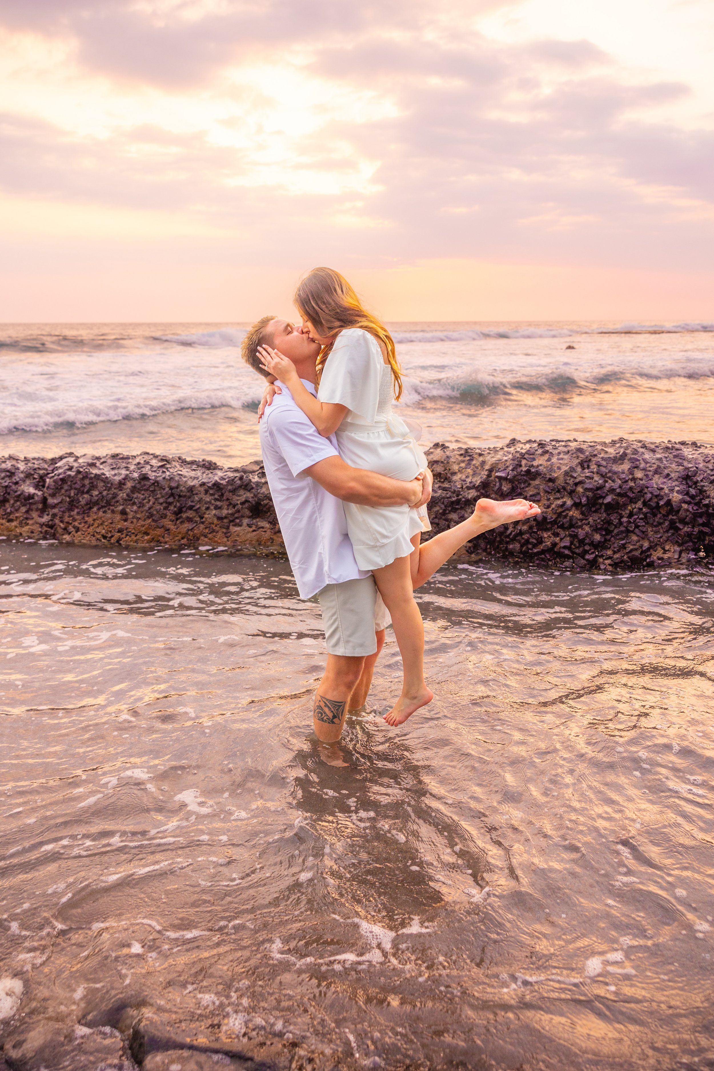 A couple embracing and kissing on the beach at sunset, with the man lifting the woman off the ground in shallow water near rocks.