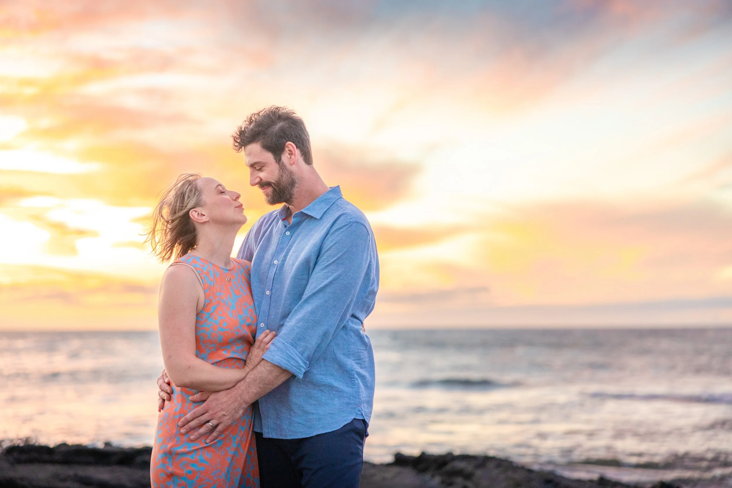 A couple standing close together on a beach at sunset, smiling and looking into each other's eyes.