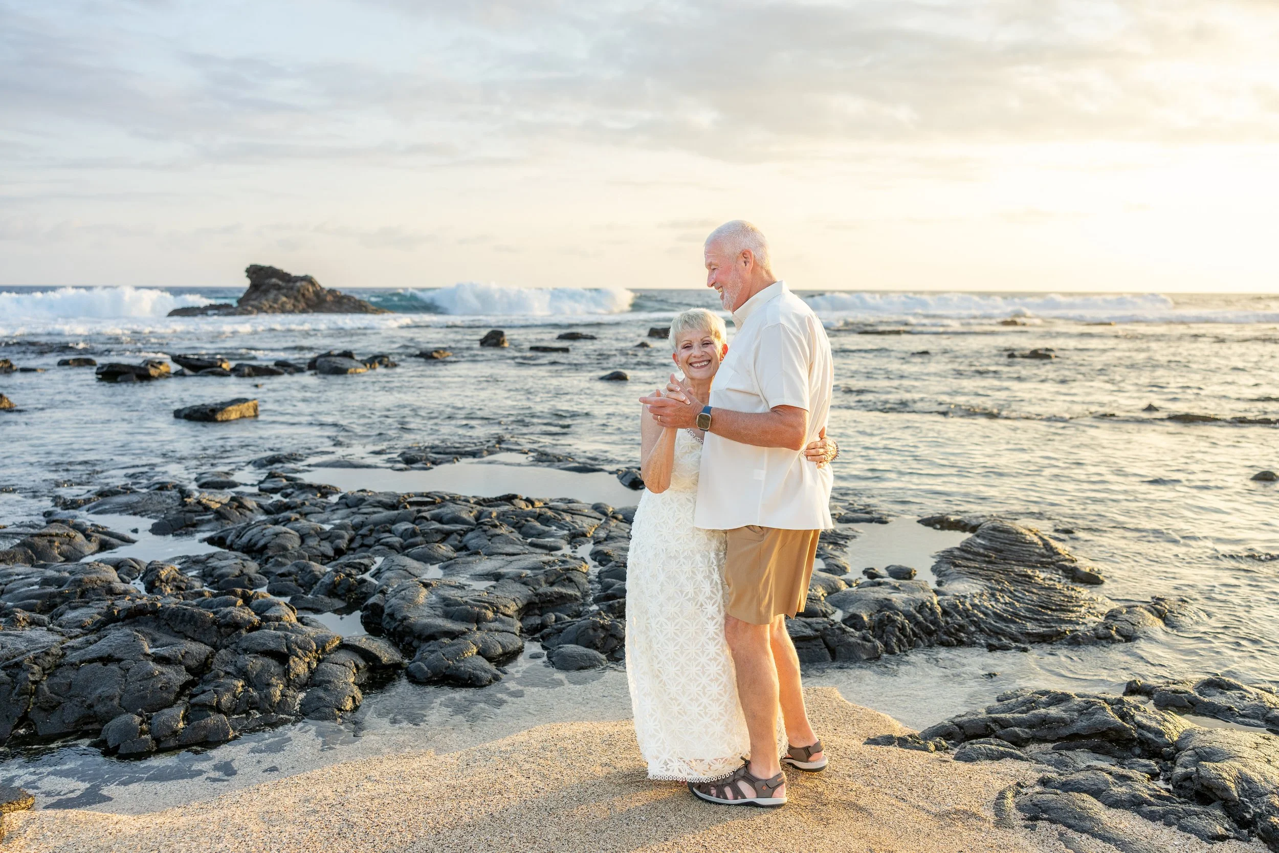 An elderly couple dancing and smiling on a rocky beach at sunset. The woman is wearing a white lace dress and the man is in a white shirt and khaki shorts.