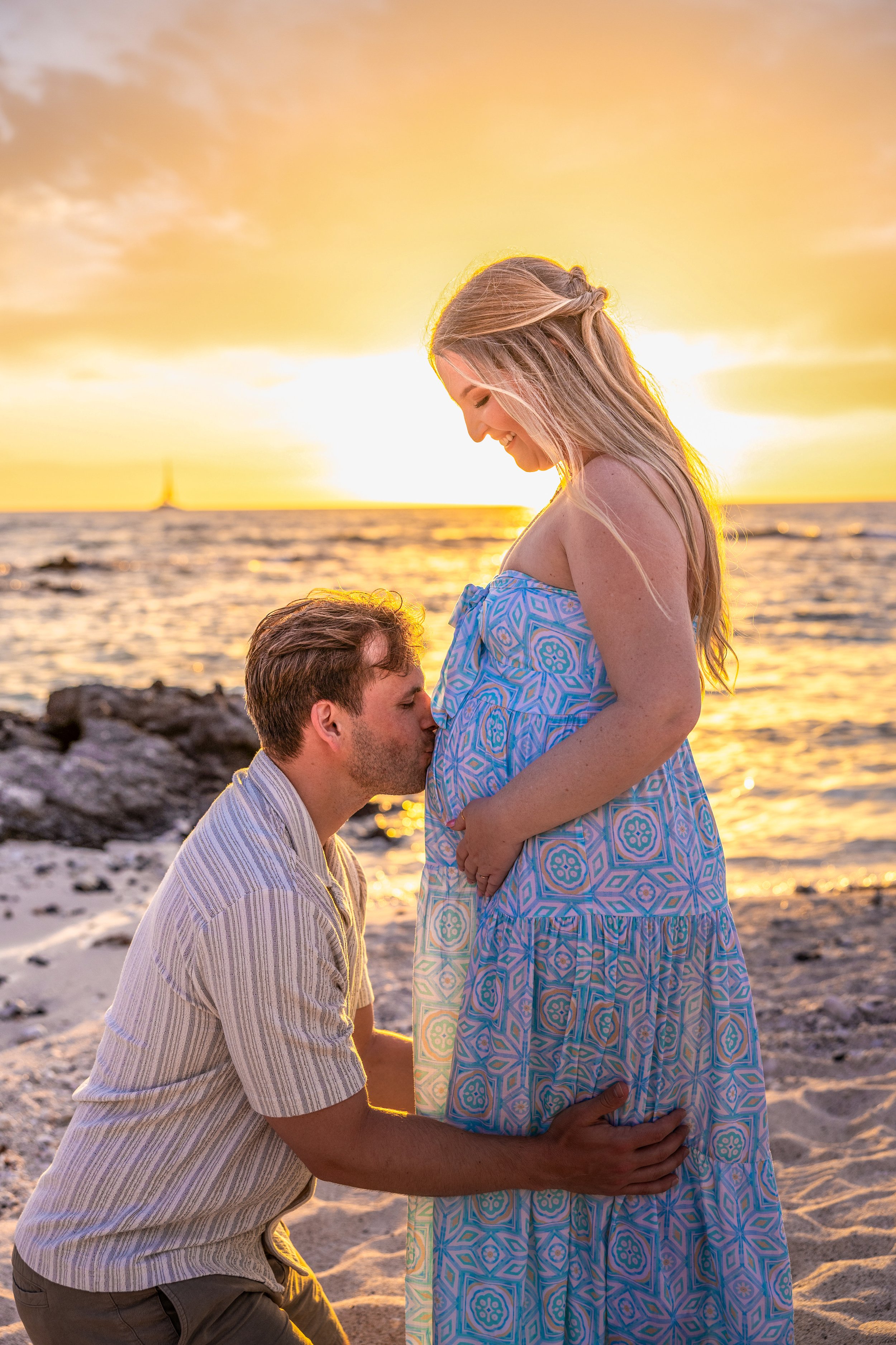 A pregnant woman in a blue patterned dress stands with a man kneeling and kissing her belly at sunset on a beach.