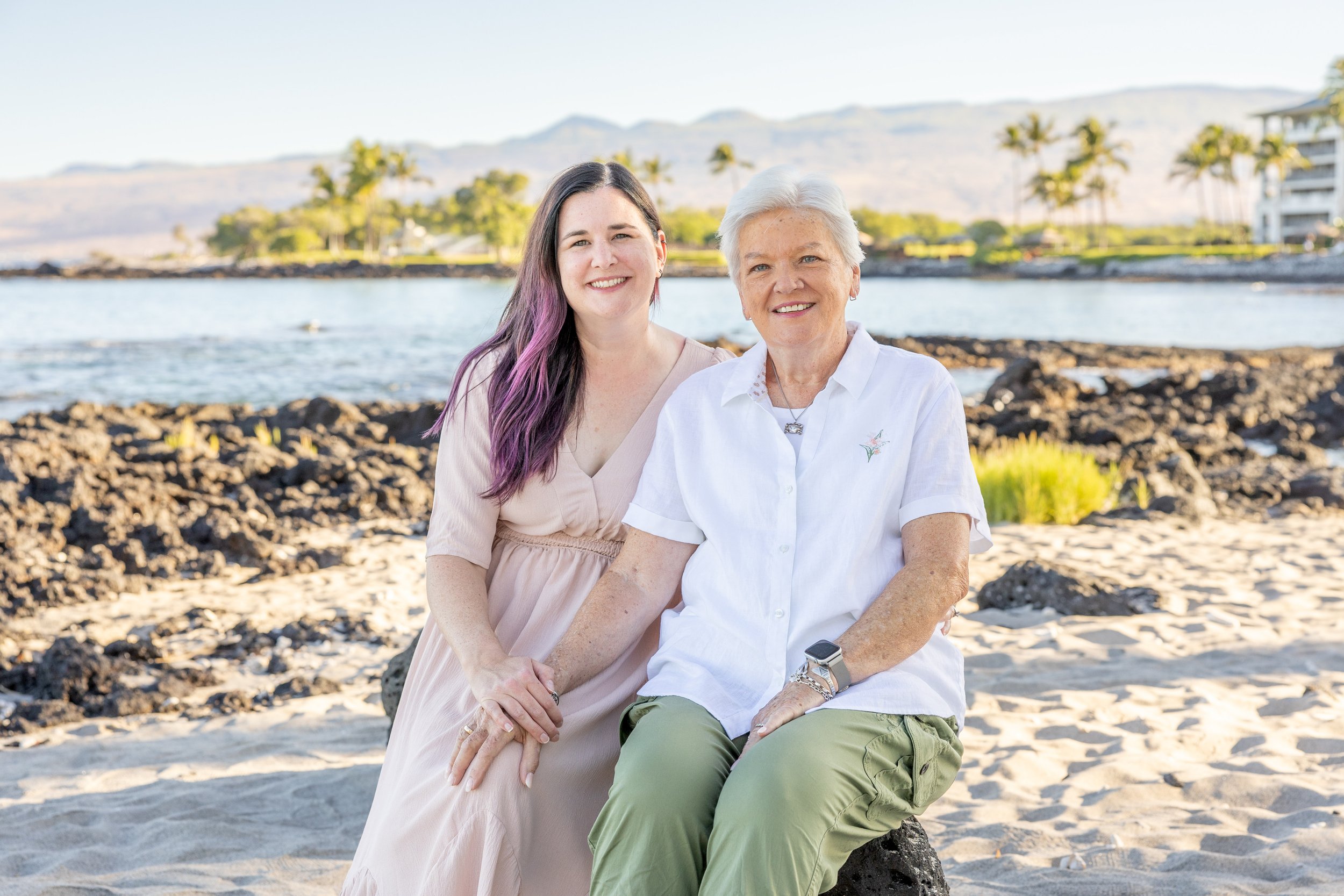 A young woman and an older woman sitting on a rock at the beach, smiling, with water and palm trees in the background.