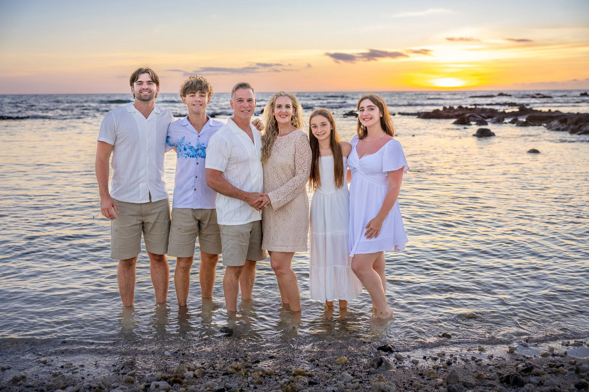 A family of six standing in ocean water at sunset, dressed in white and beige clothing, smiling at the camera.