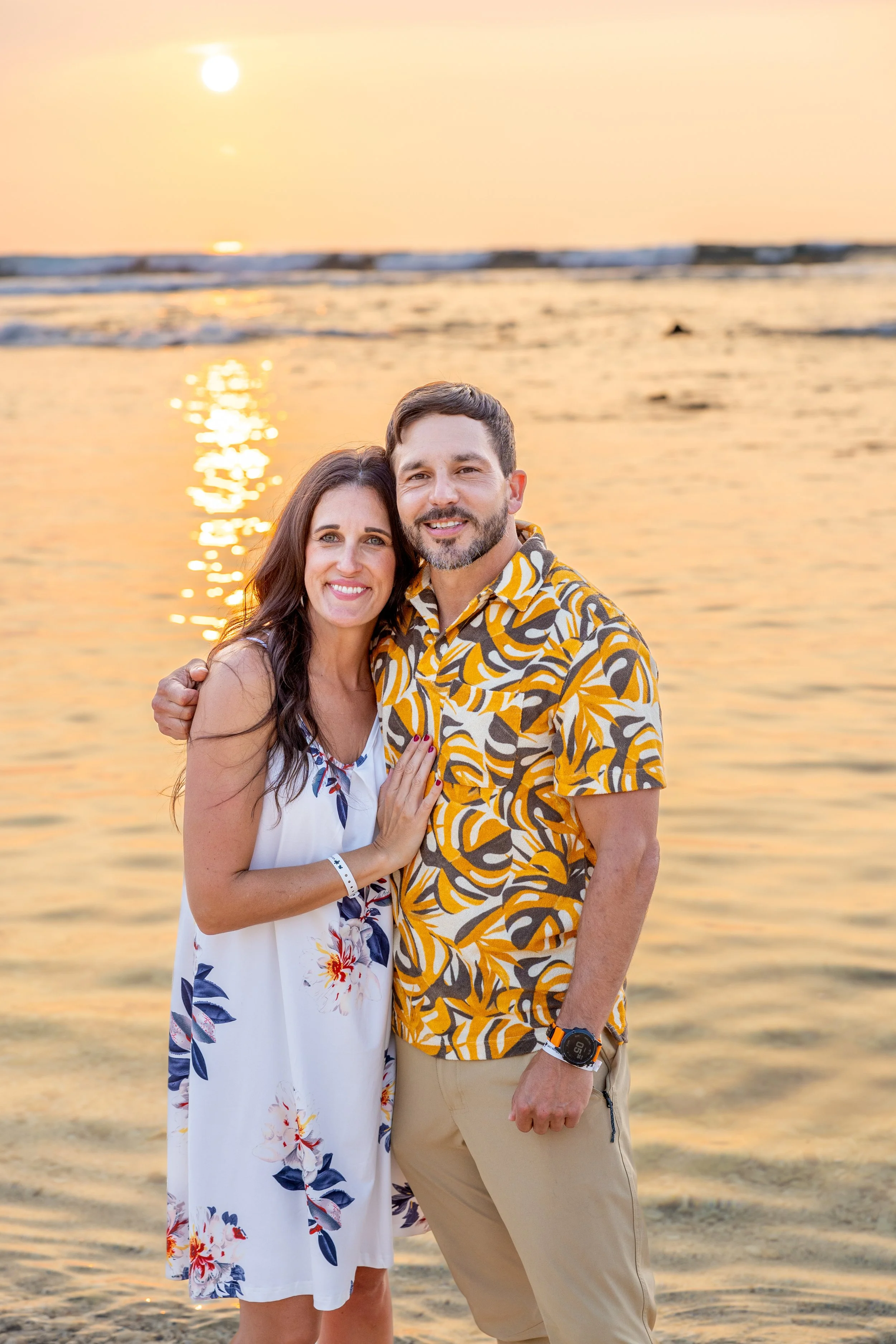 A couple smiling and embracing on a beach at sunset, with the ocean and sun reflection in the background.