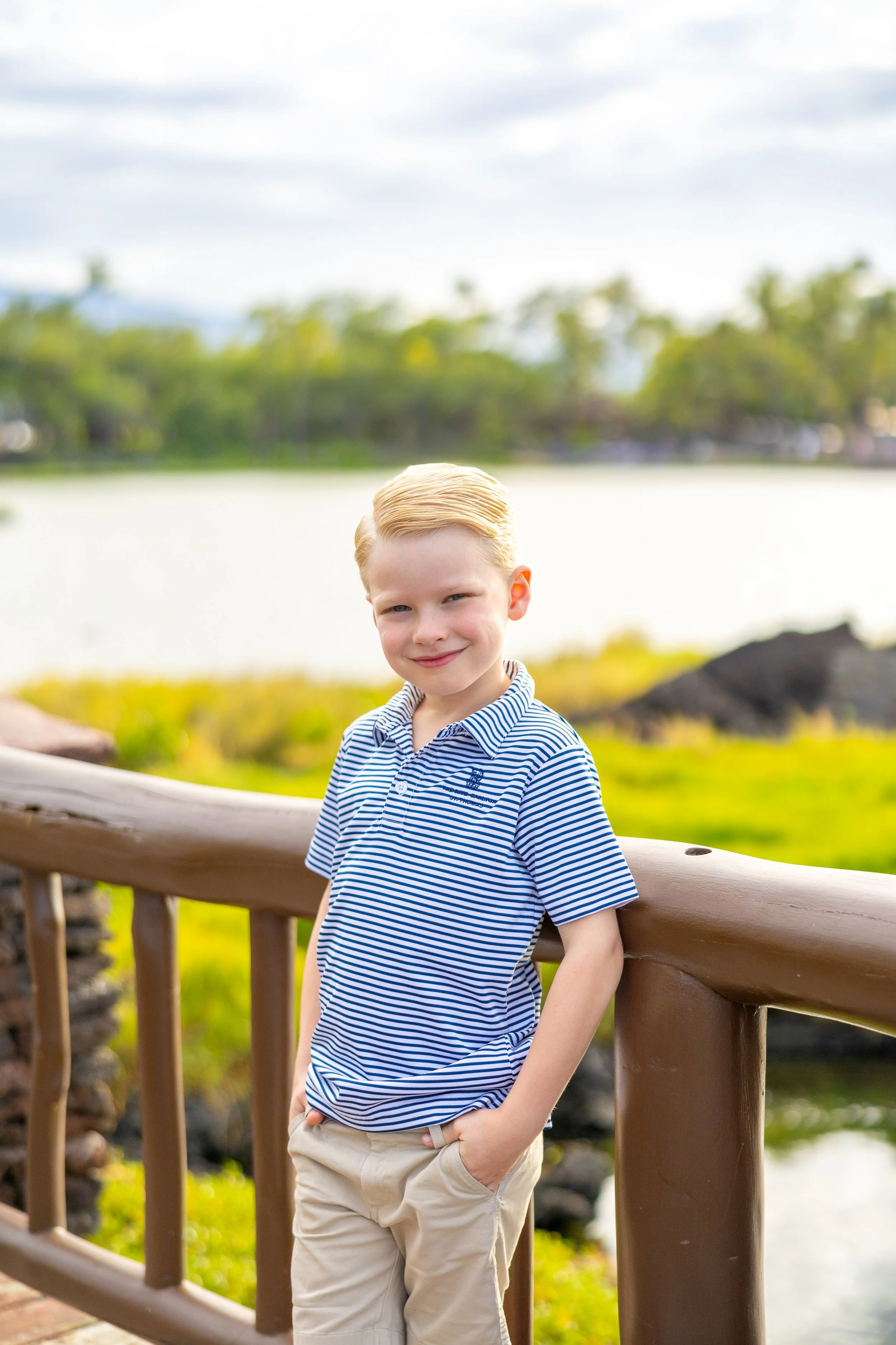 A young boy with blond hair wearing a blue and white striped polo shirt and beige pants leaning against a wooden railing outdoors with a lake, rocks, and trees in the background.