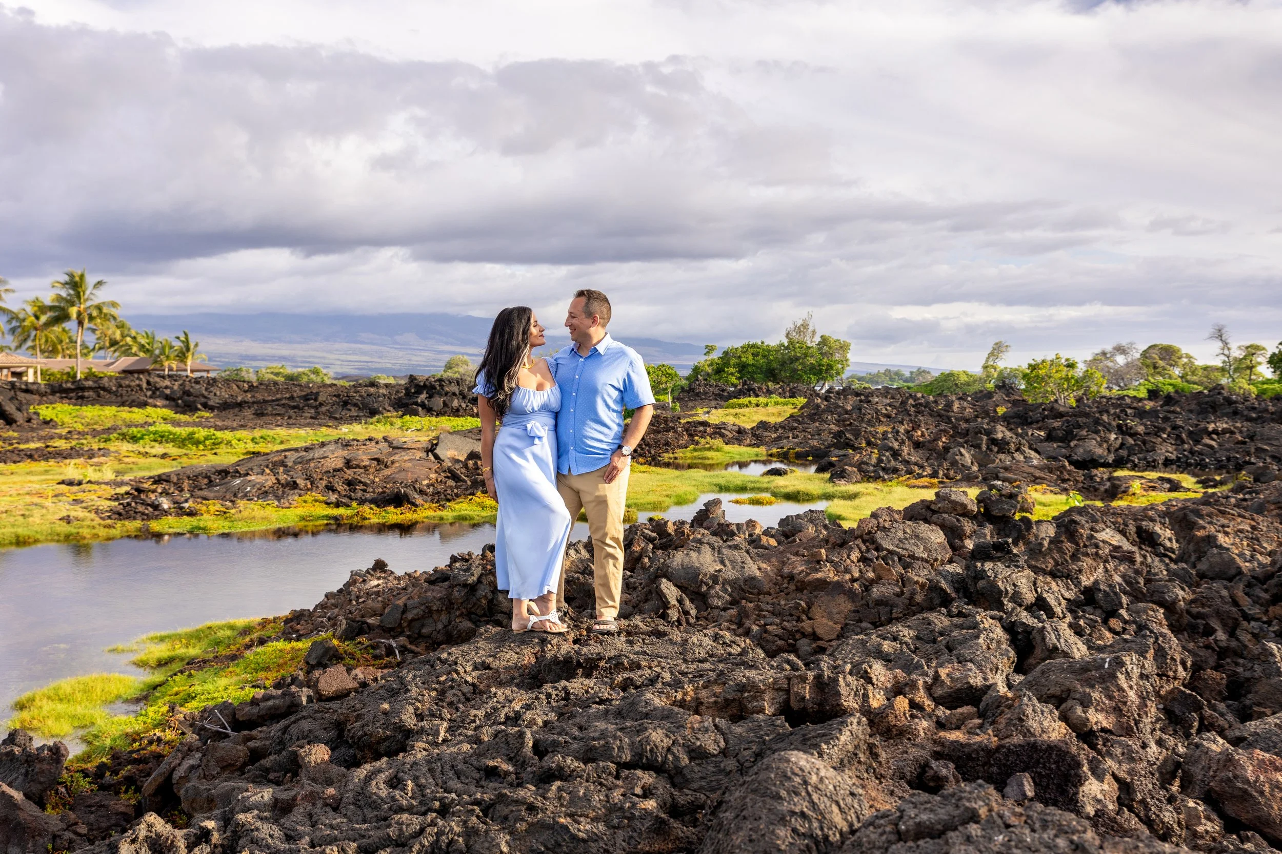 A couple standing on volcanic rocks near a small pond, with lush greenery and trees in the background, under cloudy skies.