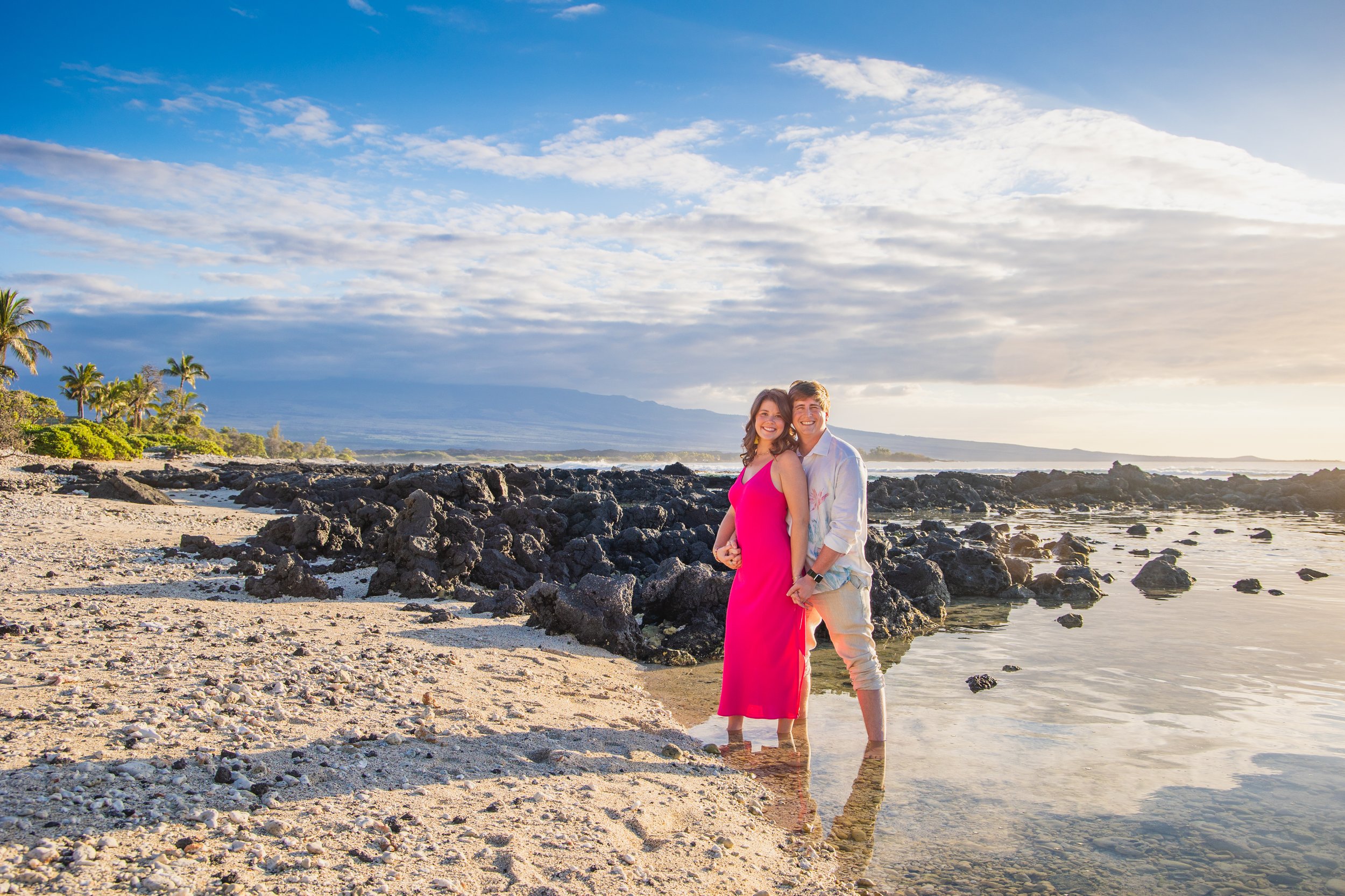 A happy couple standing on a beach during sunset, with rocks and palm trees in the background, both smiling and holding each other.