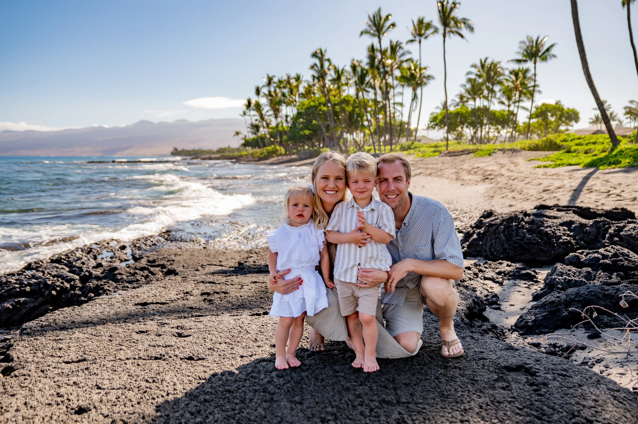 A family of four posing on a beach with palm trees and distant mountains in the background.