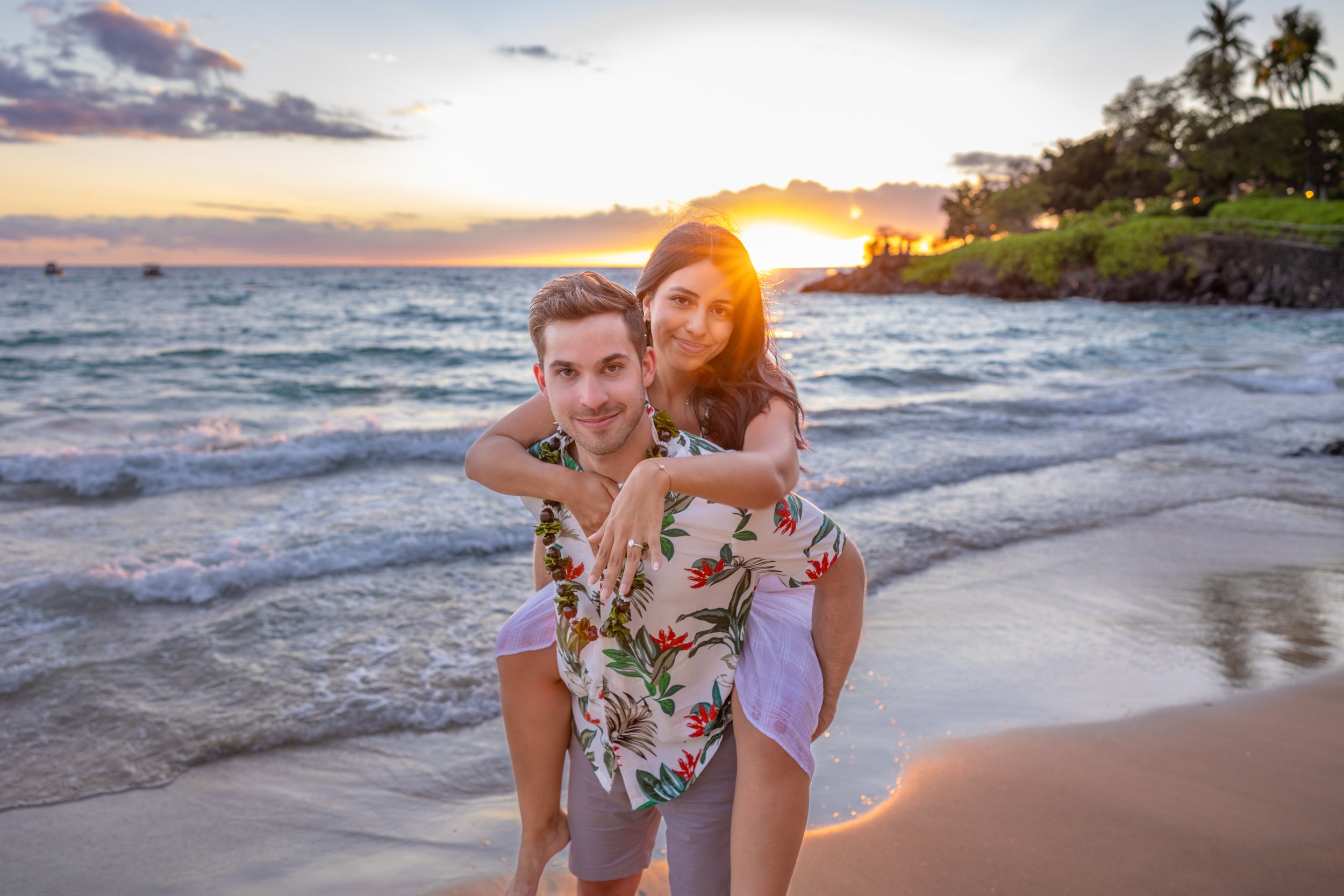 A smiling couple at the beach during sunset, with the woman giving the man a piggyback ride, in front of the ocean and a rocky shoreline with trees.