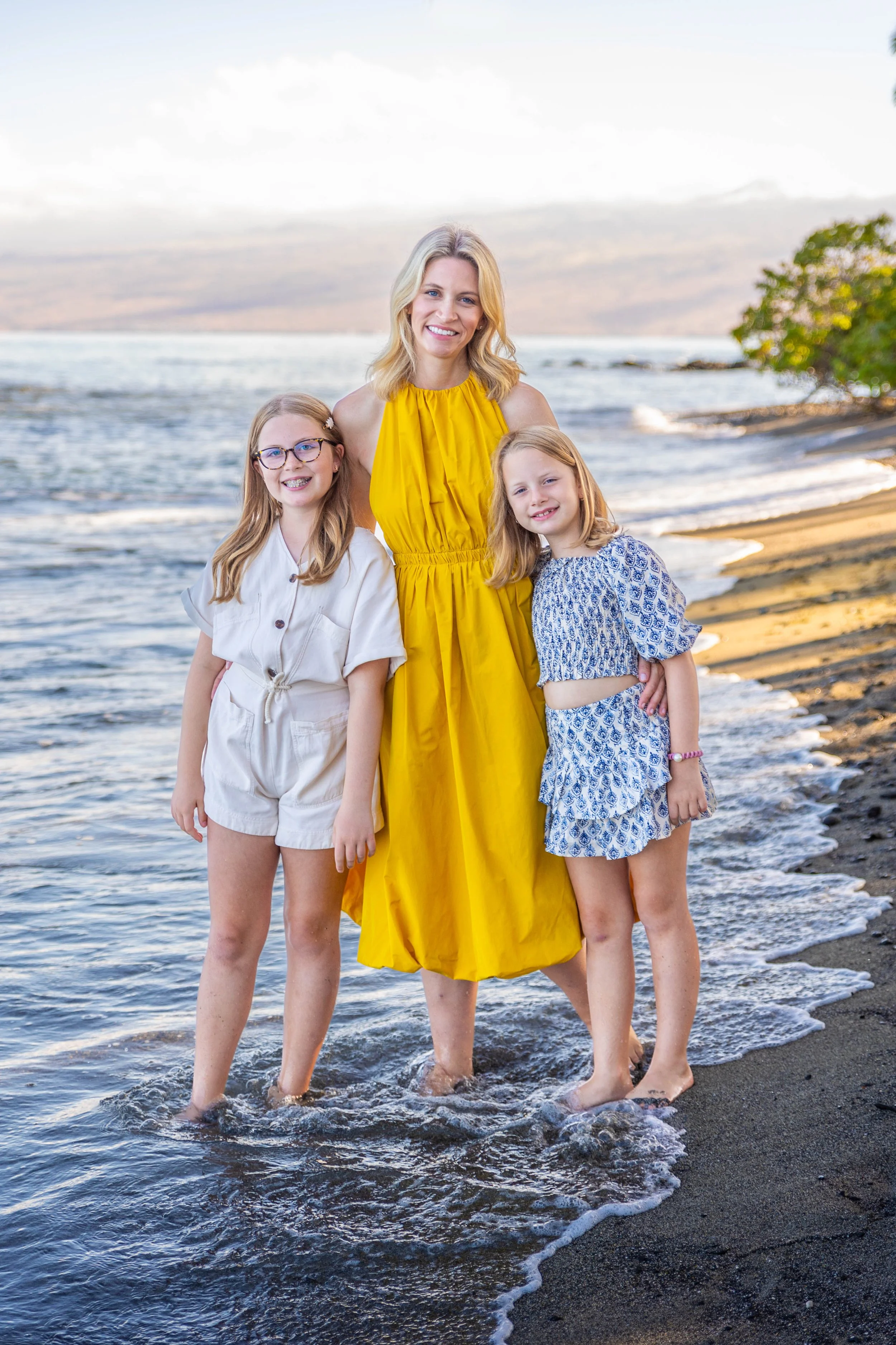 A woman in a yellow dress standing in the water at the beach with two young girls, all smiling.