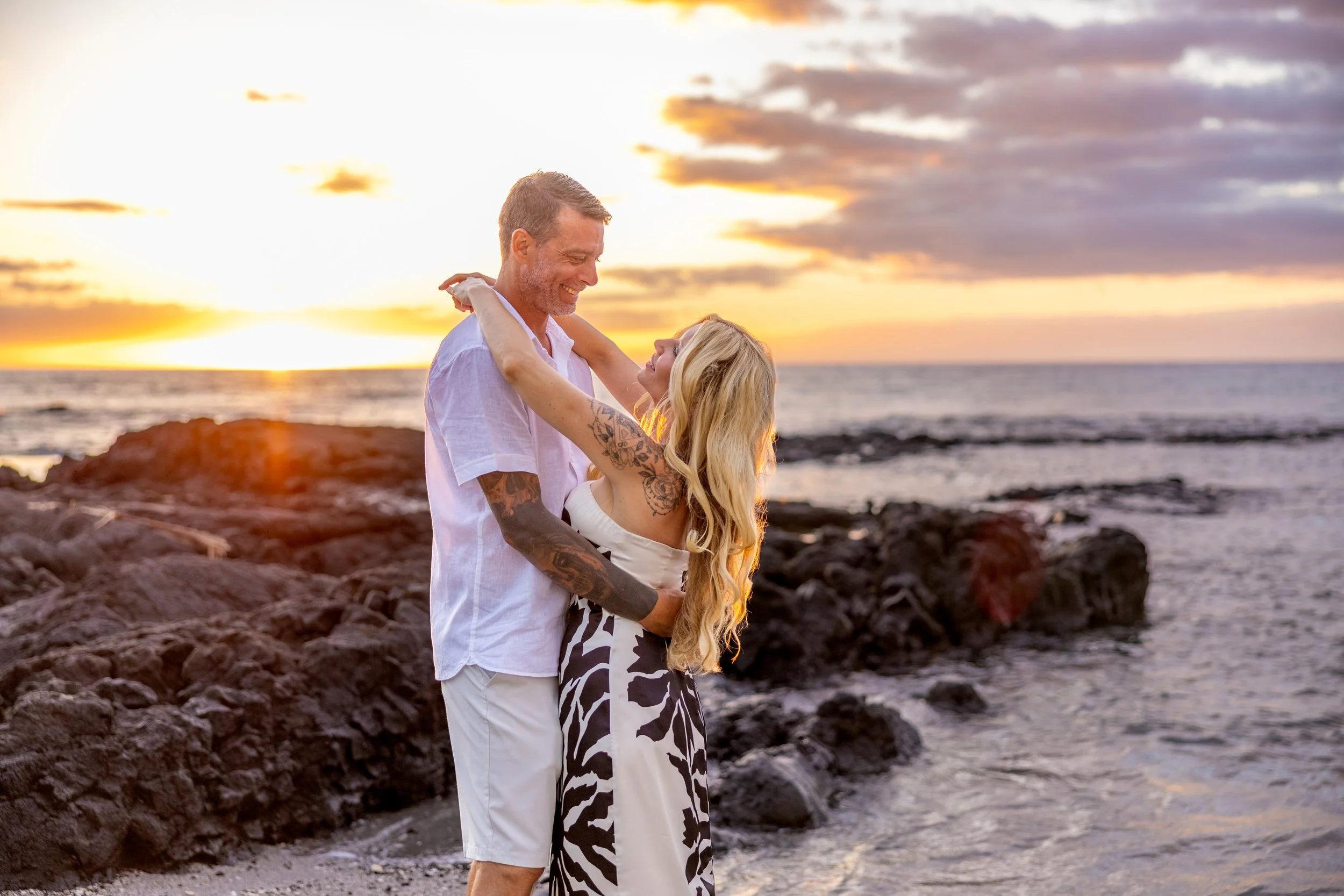 A couple dancing and smiling on a rocky beach at sunset.