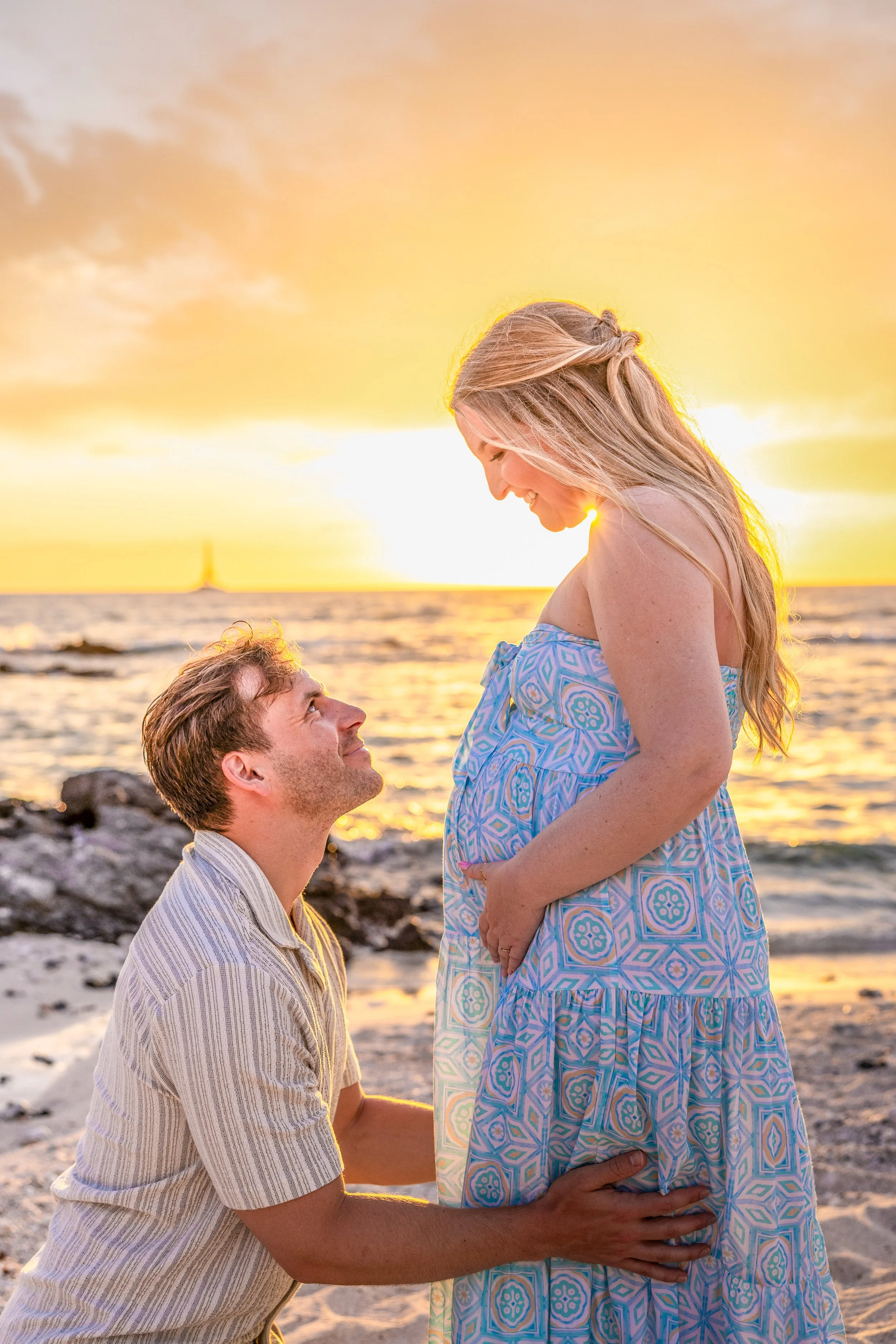 A man kneeling on the beach, holding a pregnant woman's belly, while she looks down at him during a sunset with a sailboat in the distance.