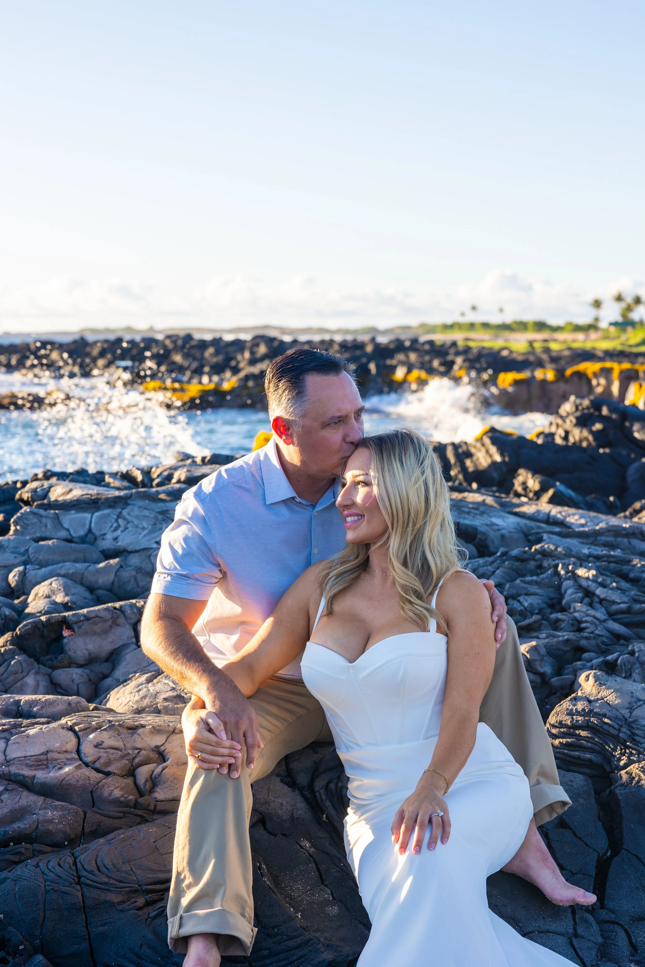 A couple sitting on rocks by the ocean, with the man kissing the woman's forehead.