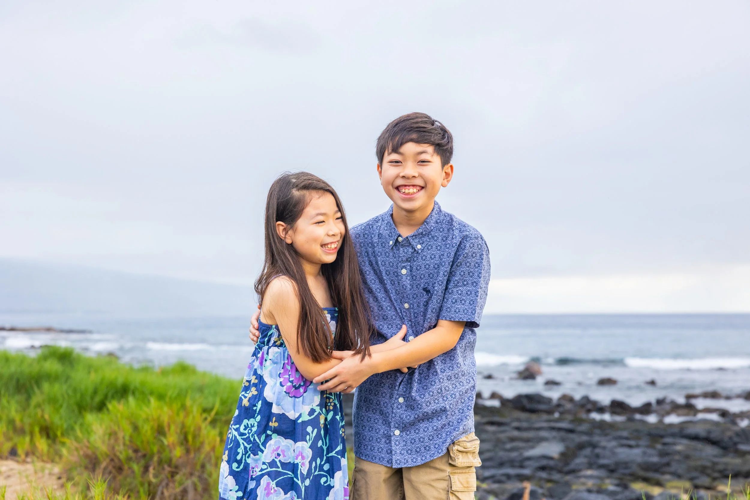 A boy and girl standing together near the ocean, smiling and hugging. The girl has long dark hair and is wearing a blue floral dress. The boy has short dark hair and is wearing a blue patterned shirt and khaki shorts.