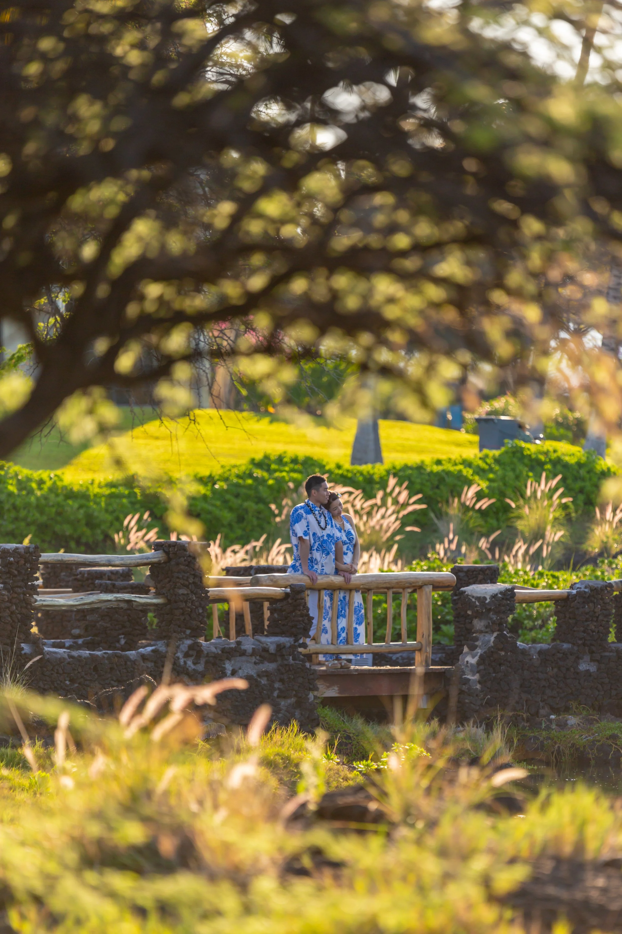 A couple in traditional floral attire standing on a small bridge in a lush park setting, viewed through foreground branches, with green grass, shrubs, trees, and sunlight in the background.
