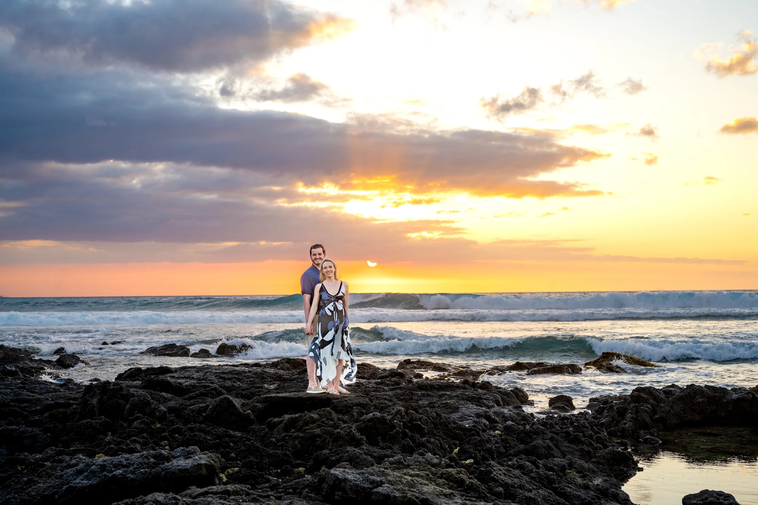 A smiling couple stands on dark volcanic rocks at the beach during sunset, with rolling waves and a colorful sky with clouds in the background.