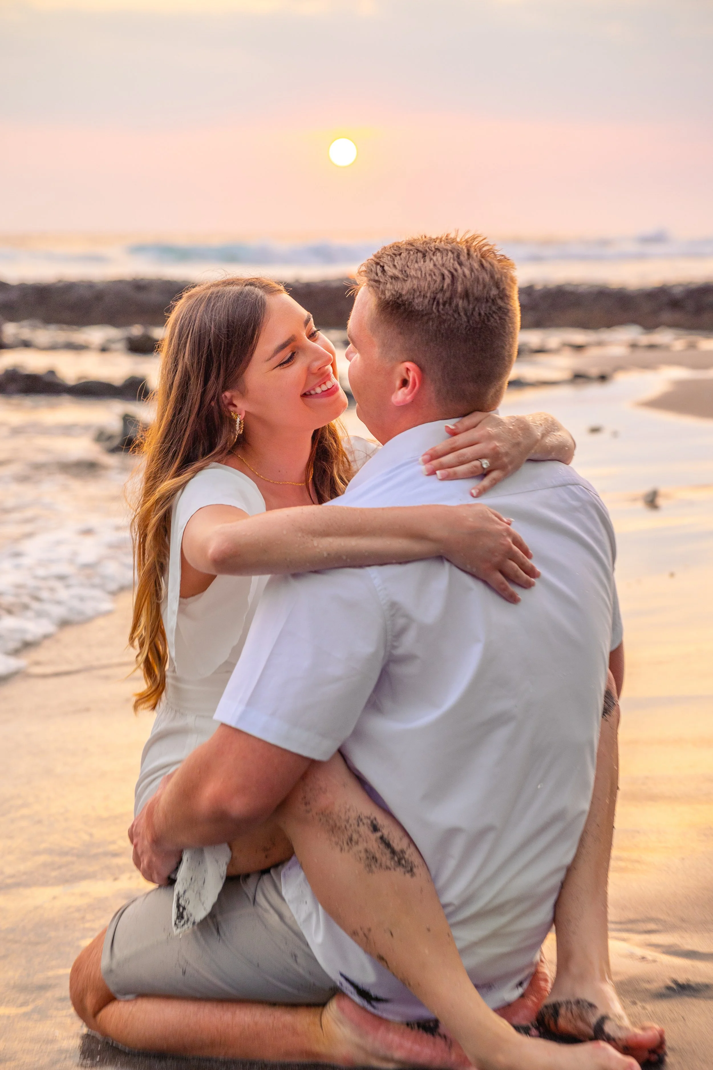 A couple embracing on a beach at sunset, with the woman sitting on the man's lap, smiling at each other.