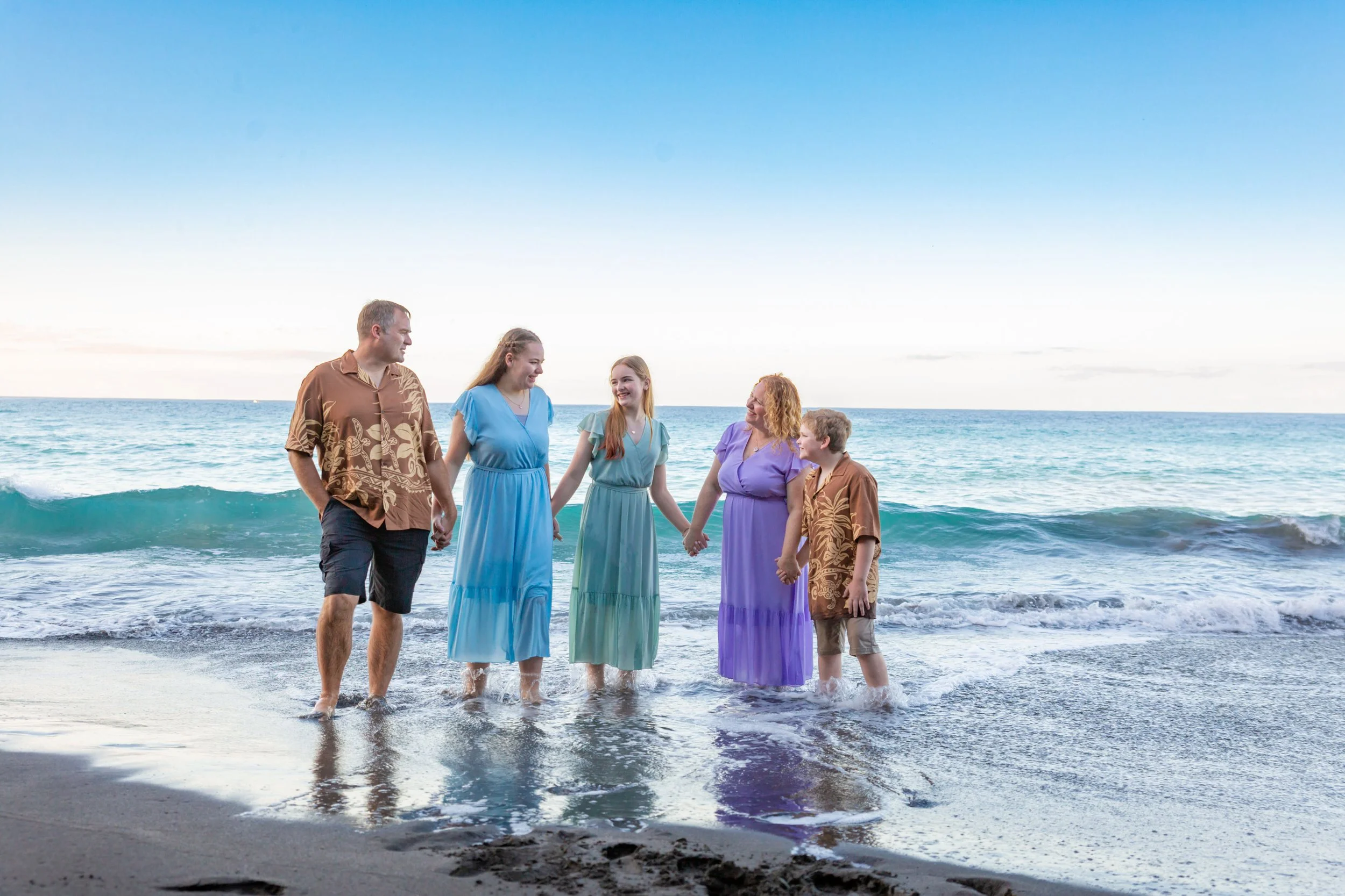 A family holding hands and standing in shallow ocean water on a beach, dressed in colorful dresses and casual clothing, with gentle waves and a clear sky in the background.