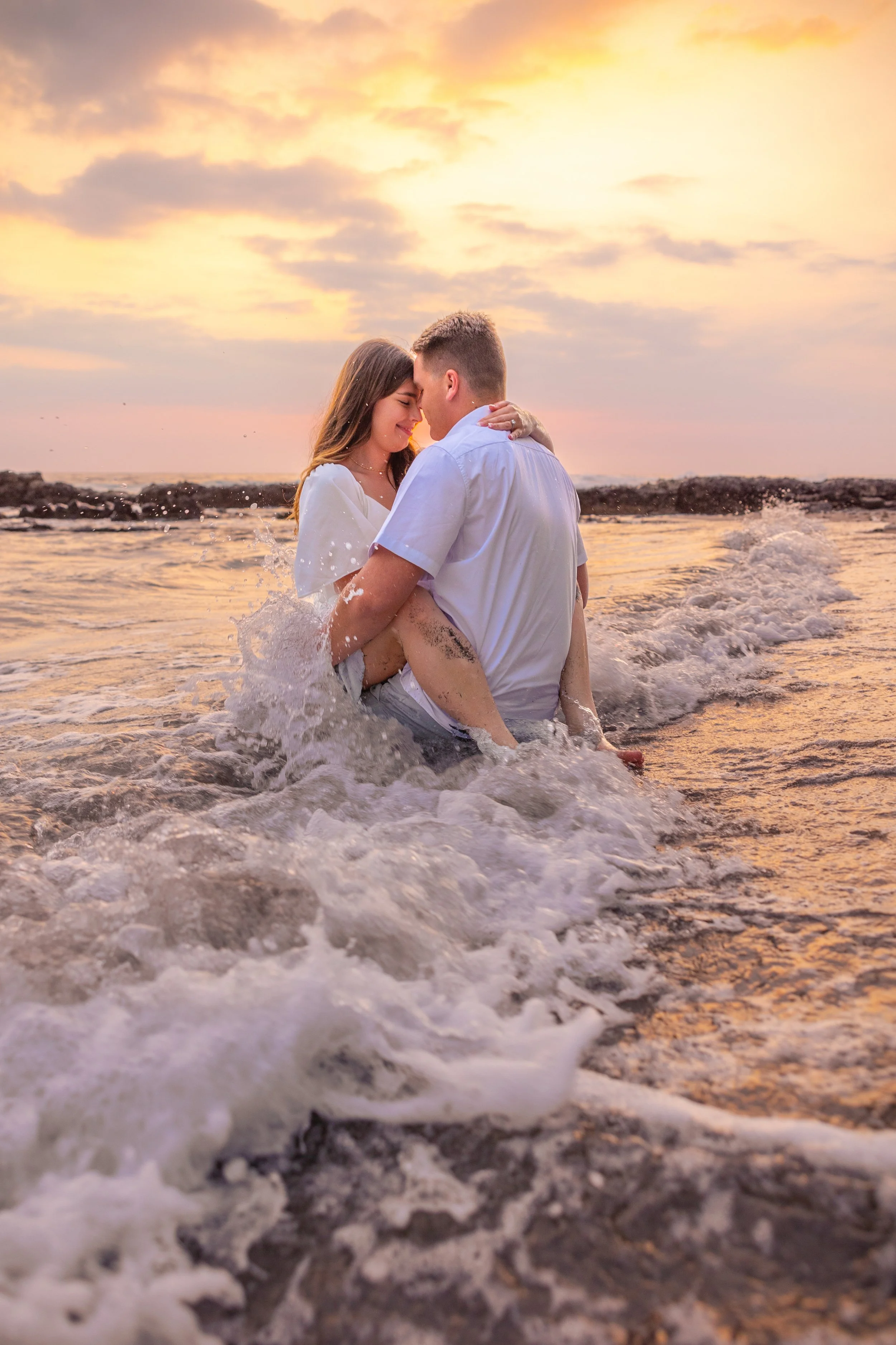 A couple embracing in the ocean at sunset, with waves crashing around them and a colorful sky overhead.