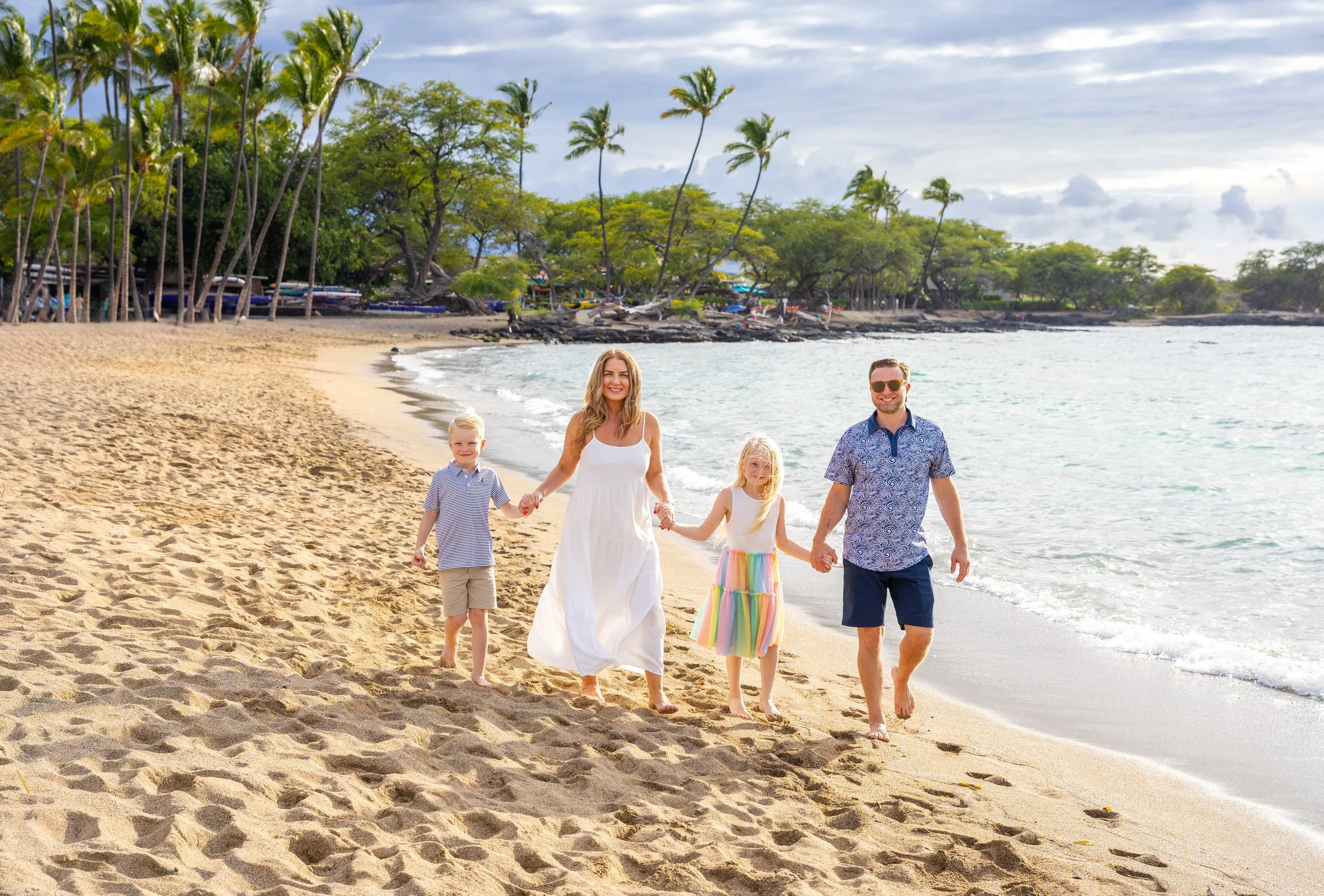 A family of four walking hand in hand on a sandy beach near the ocean, with palm trees and lush greenery in the background.