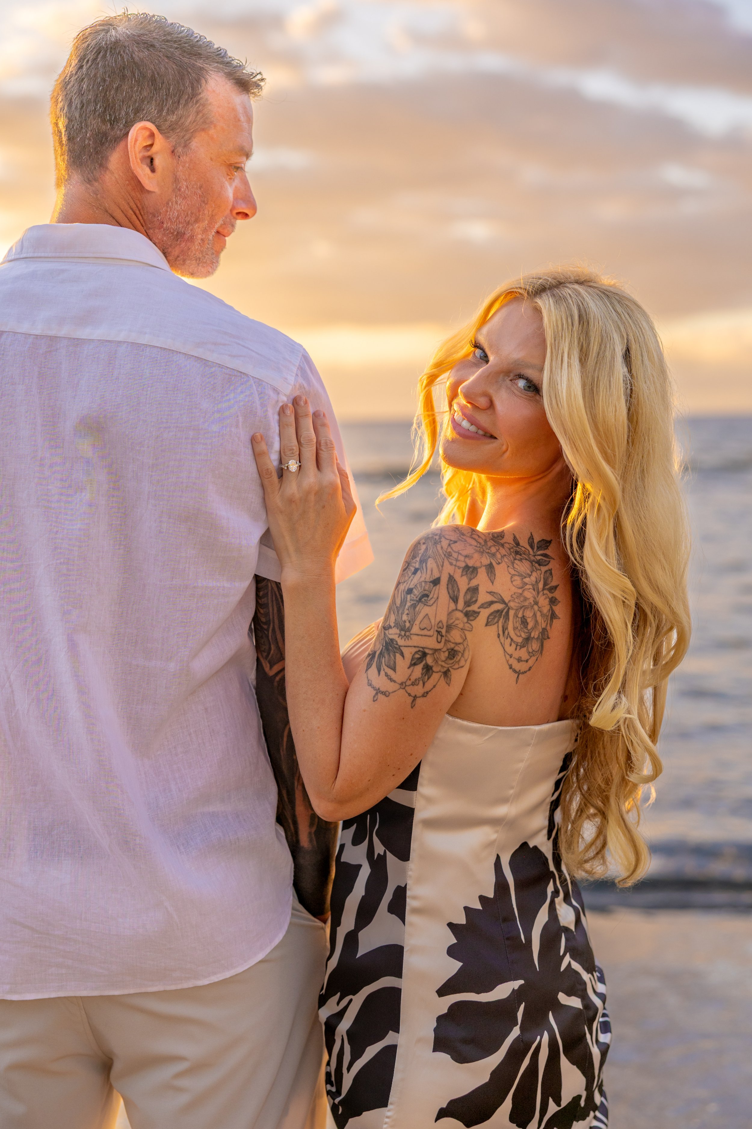 A man and a woman standing close together at the beach during sunset, with the woman showing her engagement ring.