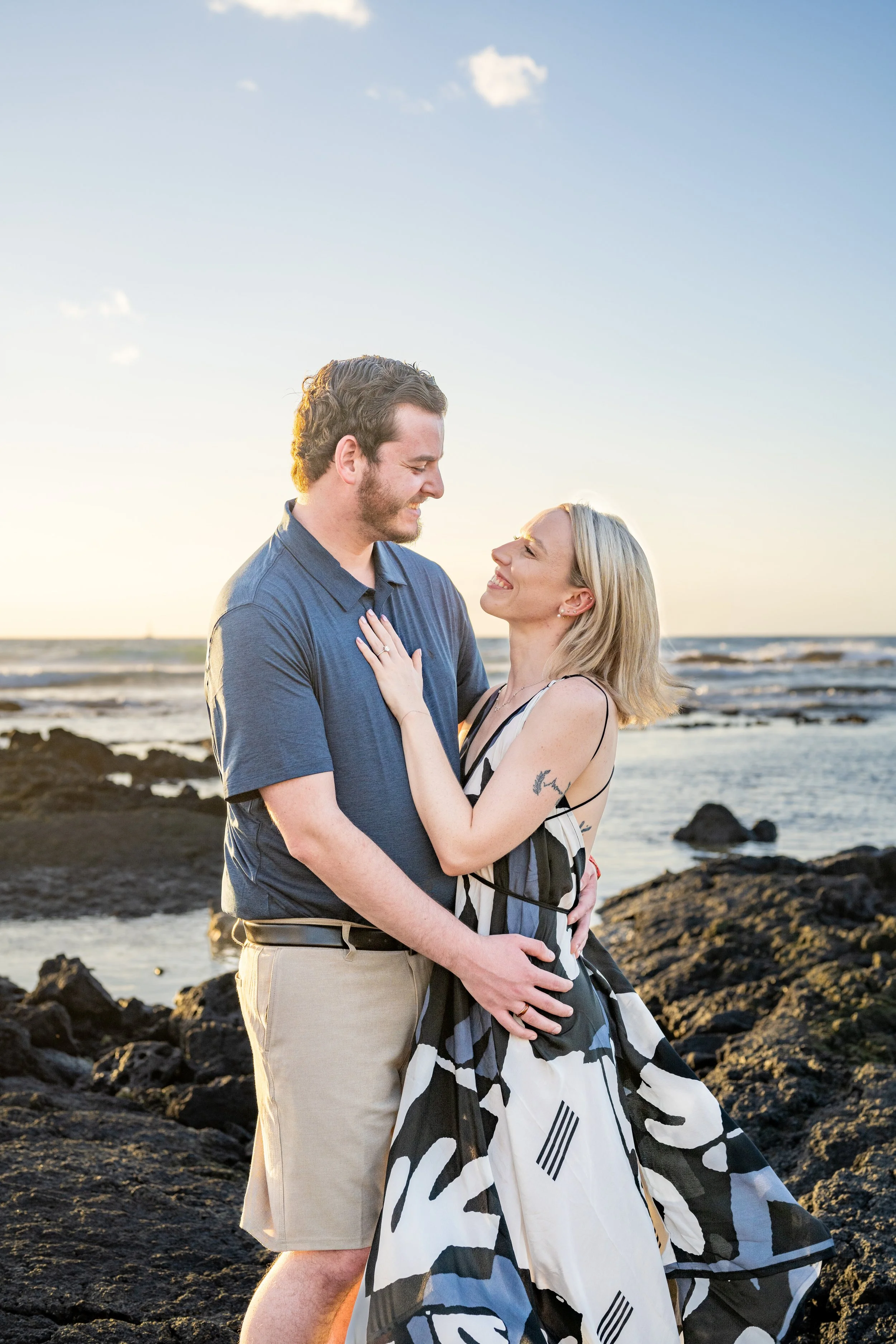 A couple standing on rocky shoreline at sunset, smiling and looking into each other's eyes.