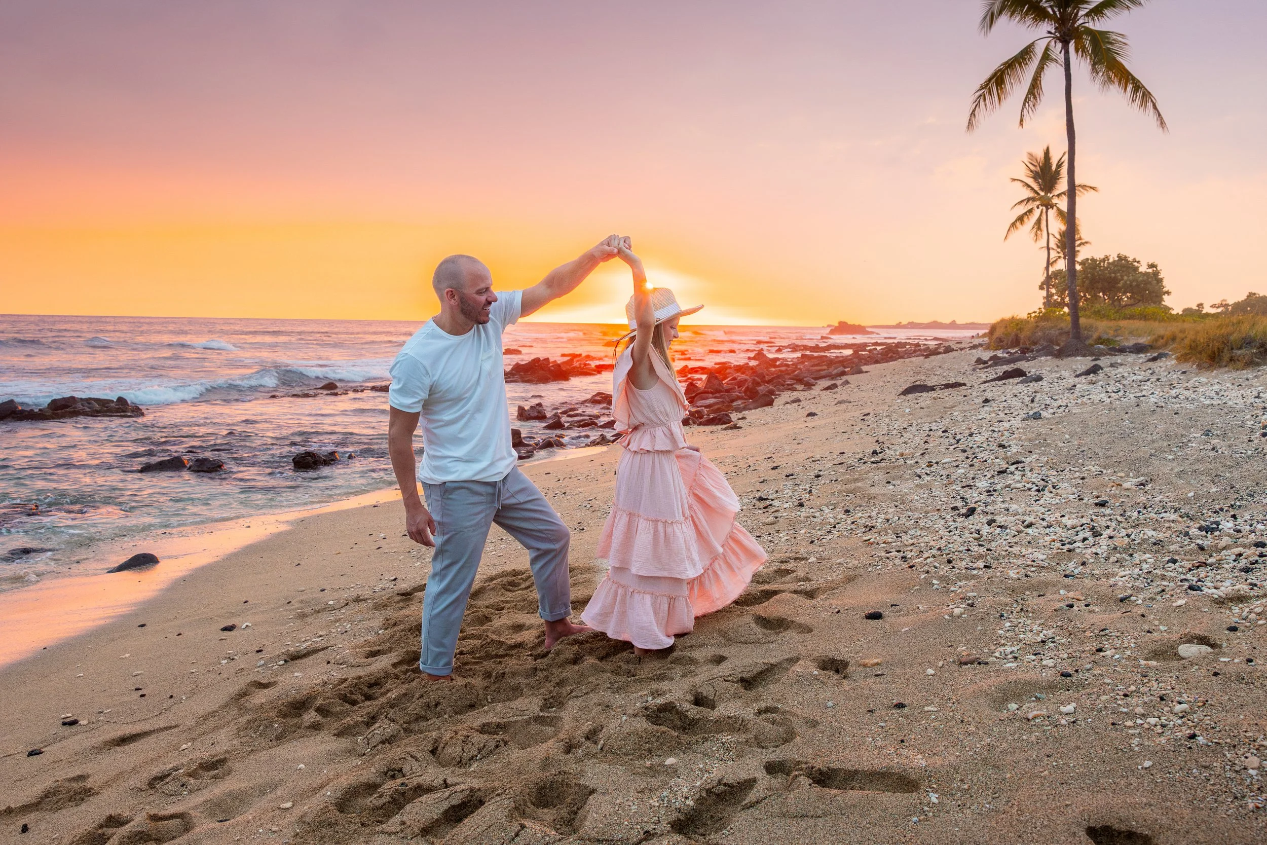 A man and woman on a beach at sunset, with the man spinning the woman around, both smiling. The woman is wearing a pink dress and a sun hat, and the man is in a white shirt and gray pants. There are two palm trees and rocks along the shoreline.