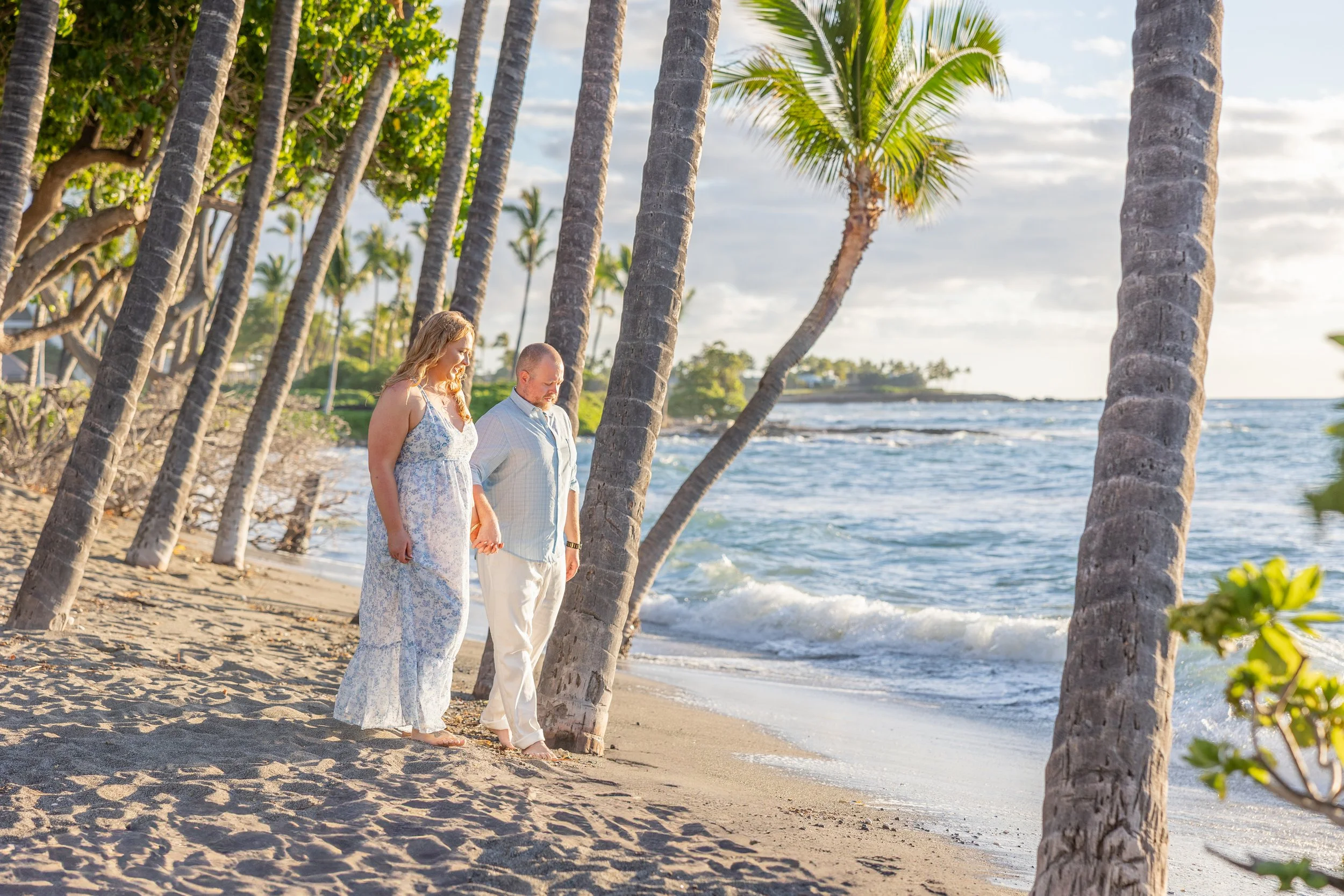 A couple holding hands and walking along a tropical beach lined with palm trees, with the ocean waves in the background.