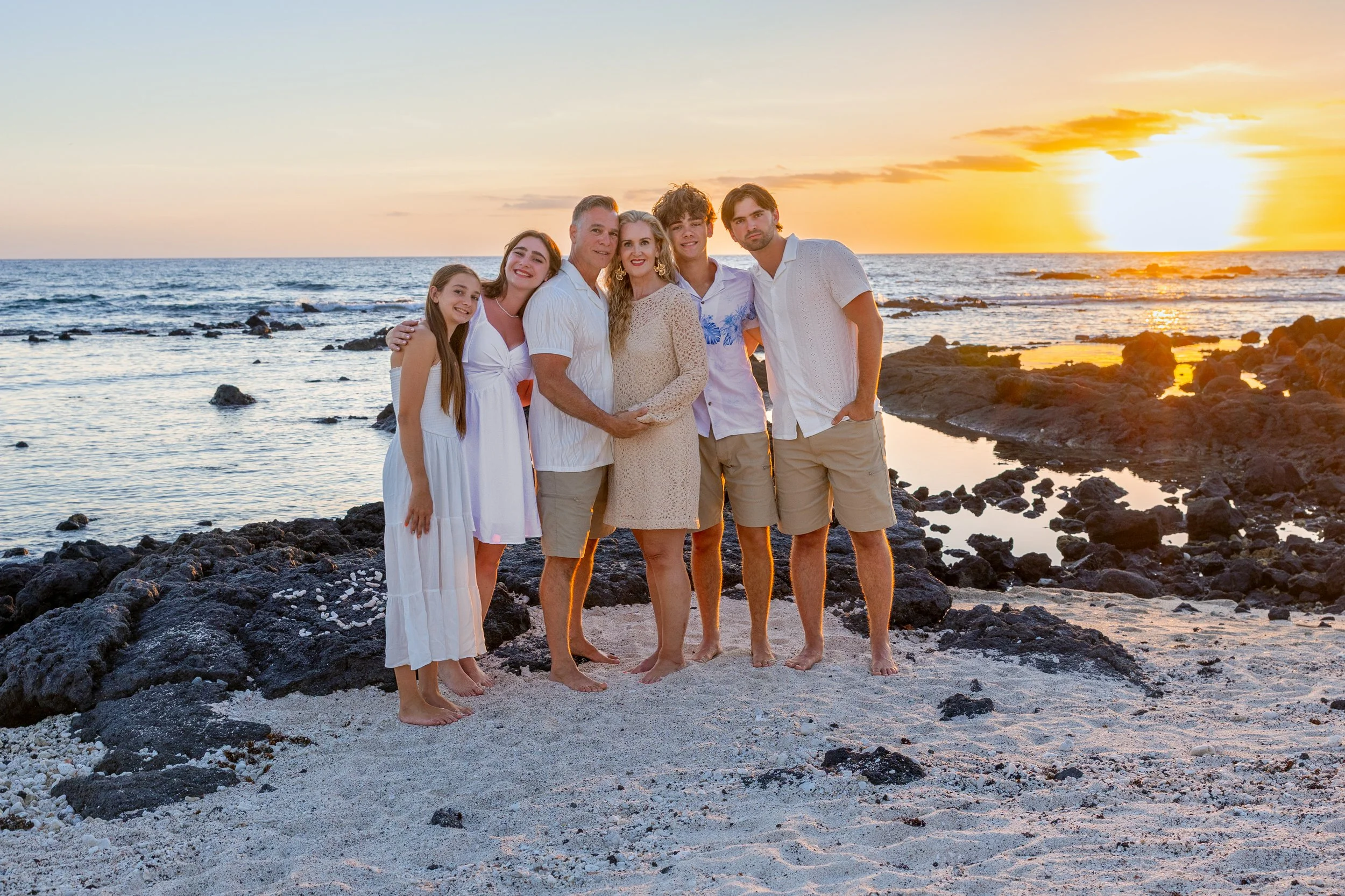 Family of seven standing on rocky beach at sunset, smiling and embracing, wearing light-colored clothing.