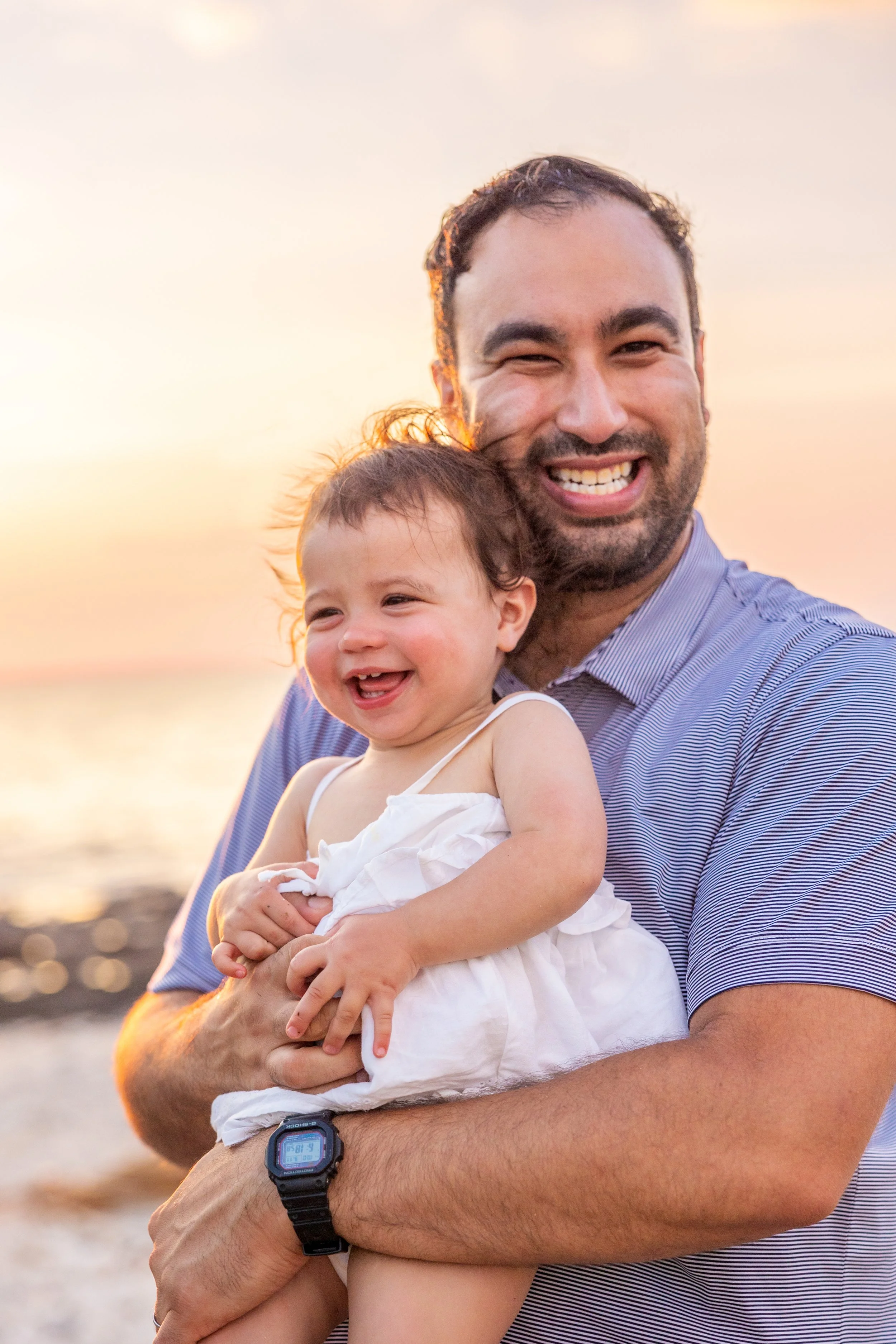 A man holding a smiling toddler girl at the beach during sunset.
