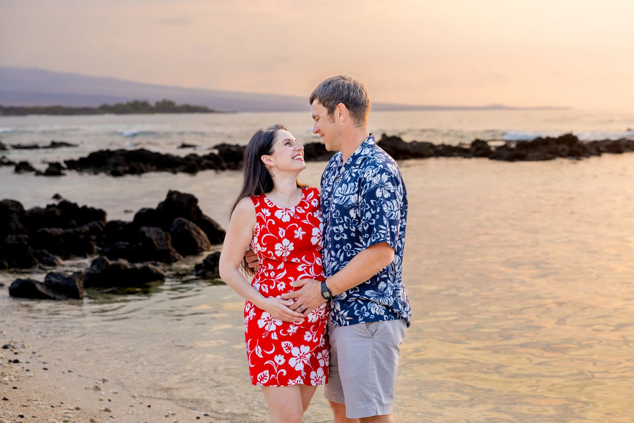 A couple standing on a beach during sunset, looking at each other and smiling. The woman is wearing a red dress with white floral patterns, and the man is wearing a blue Hawaiian shirt with beige shorts.