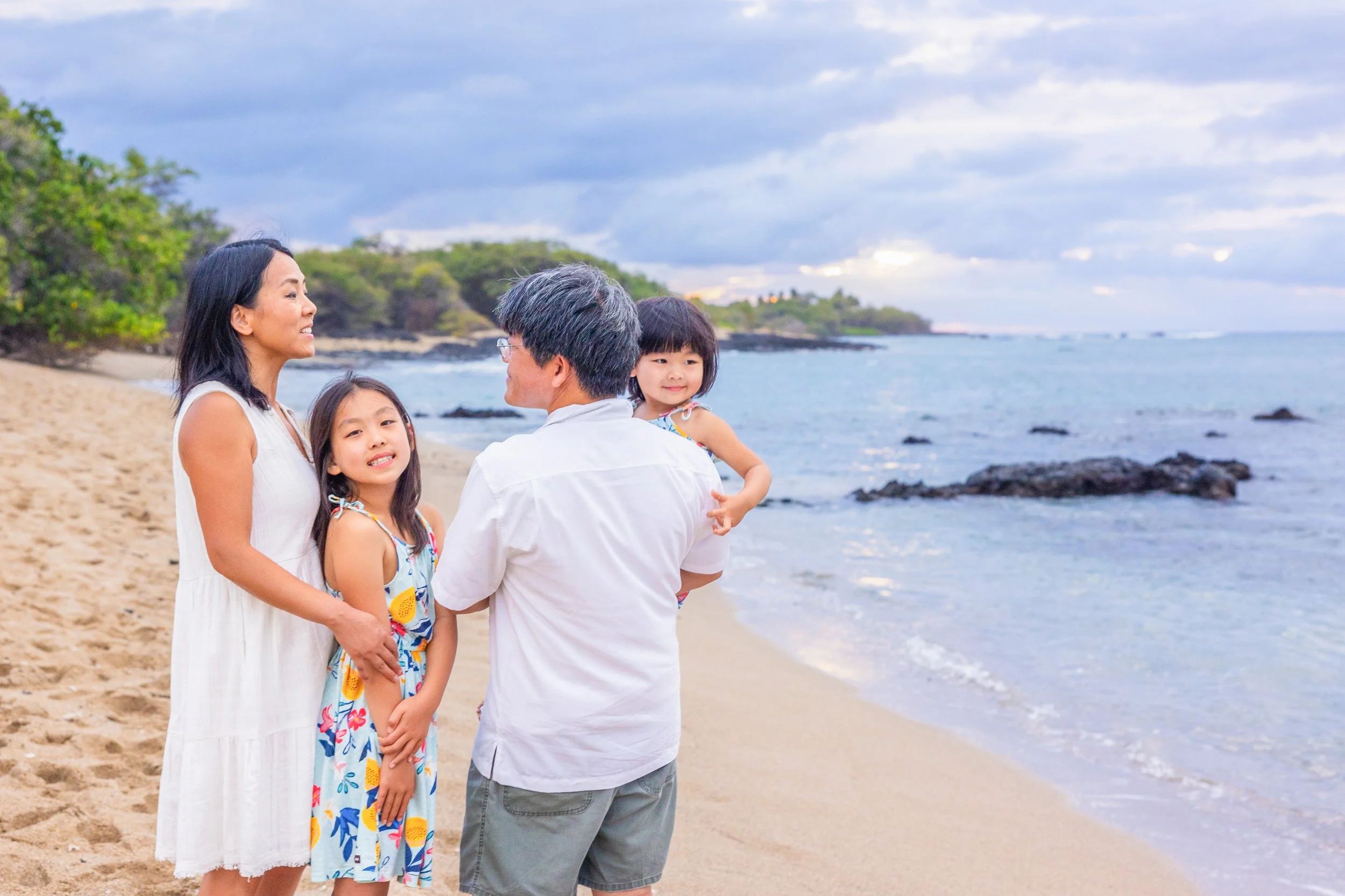 A family of four stands on a beach, facing each other and smiling. The mother and daughter are on the left, and the father is holding the younger daughter on the right. The sky is partly cloudy, and the ocean waves gently wash up on the sandy shore w