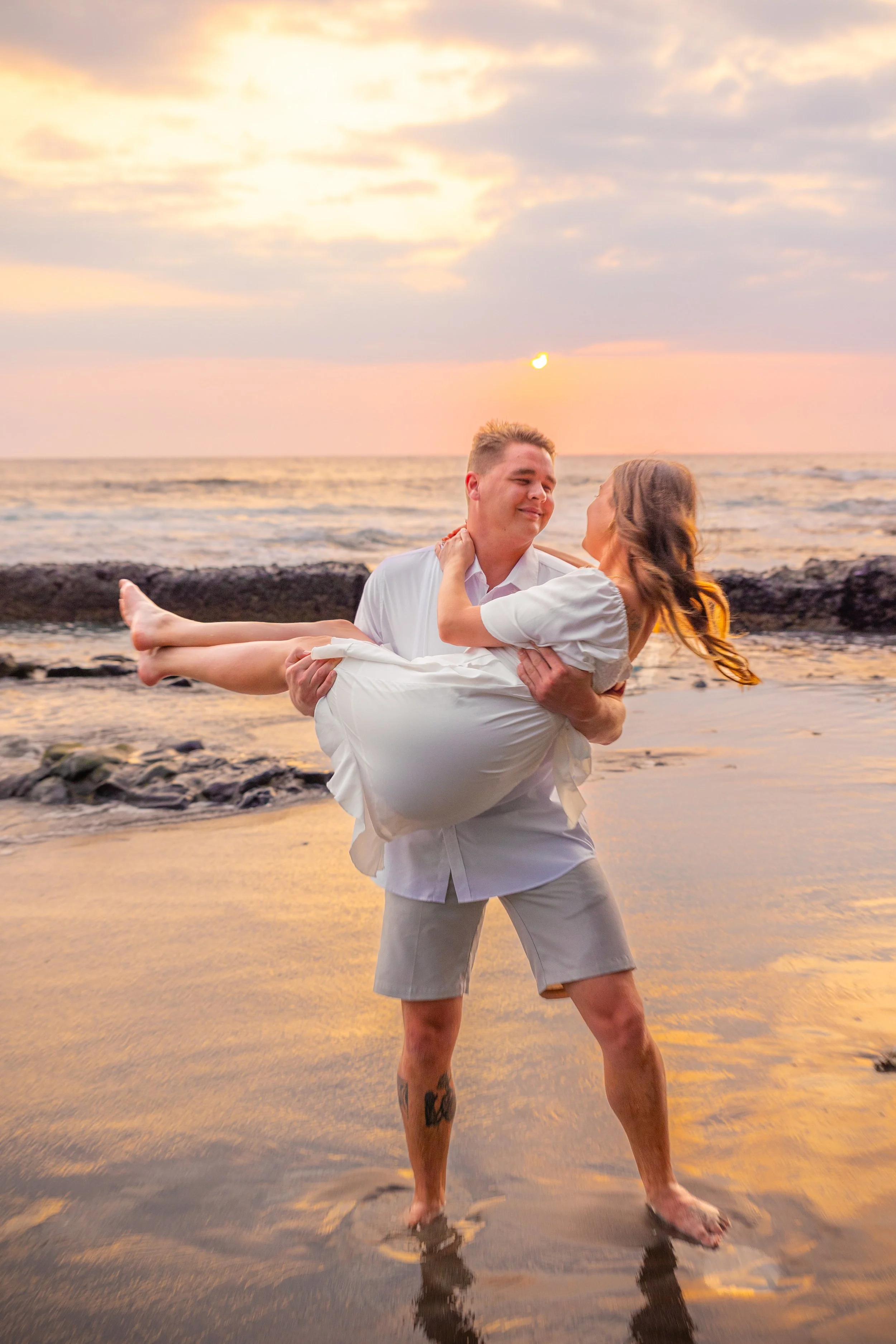 A man holding a woman in a white dress in his arms on the beach at sunset, with the ocean and sky in the background.