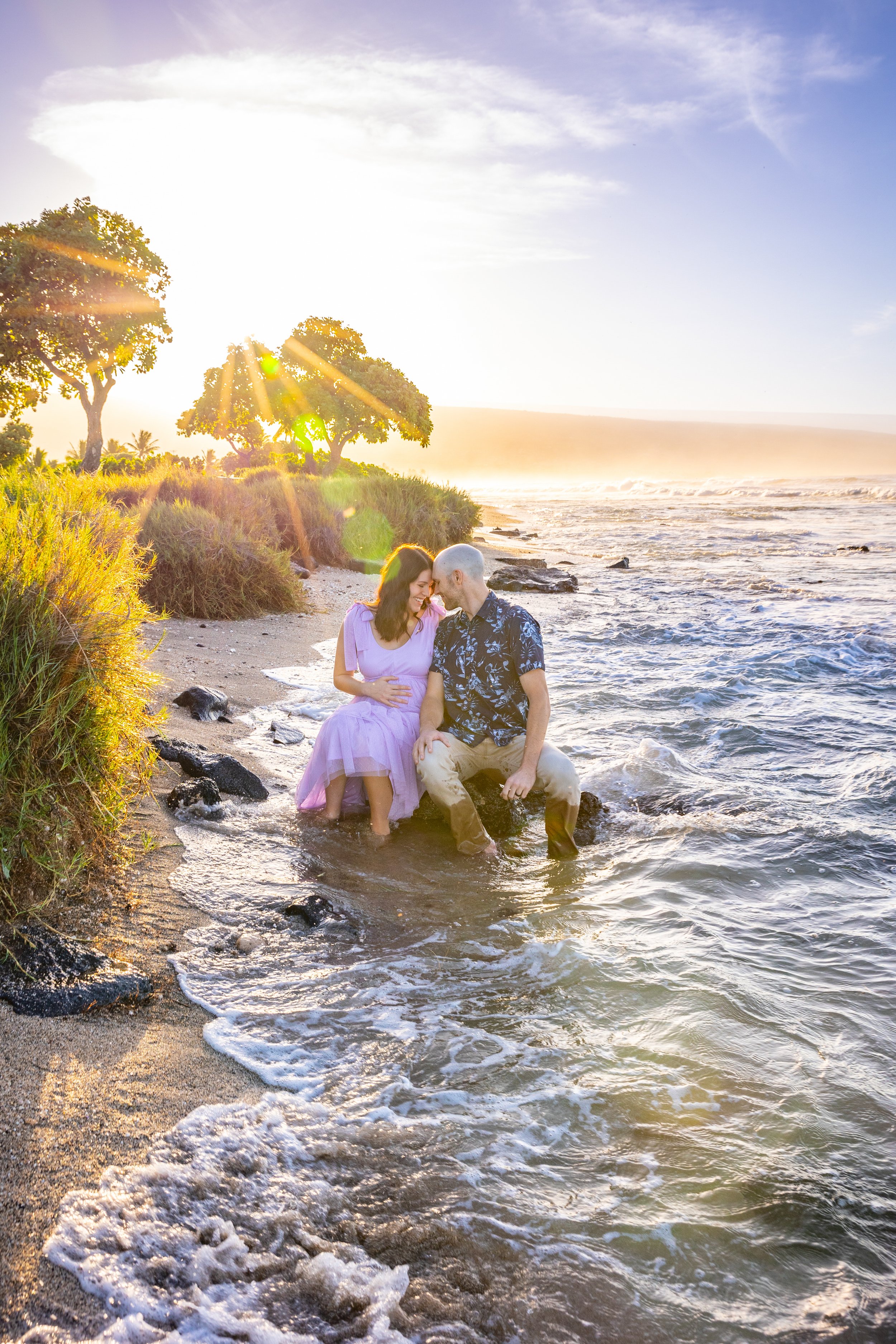 A couple sitting on the beach in the shallow water at sunset, embracing and smiling, surrounded by trees and ocean waves.