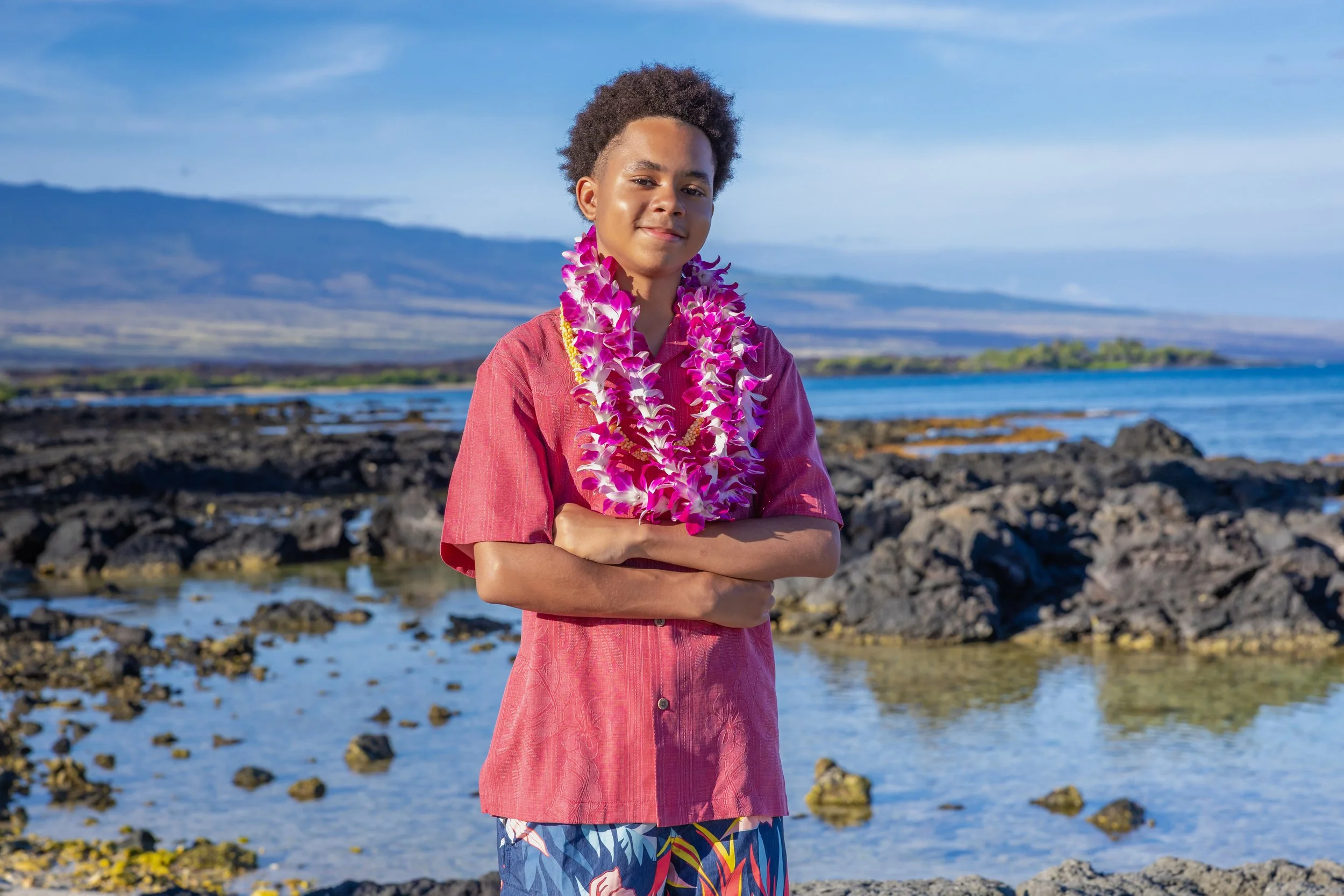 A young boy with curly hair wearing a pink shirt, patterned shorts, and a purple lei, standing on a rocky shoreline with the ocean and mountains in the background.