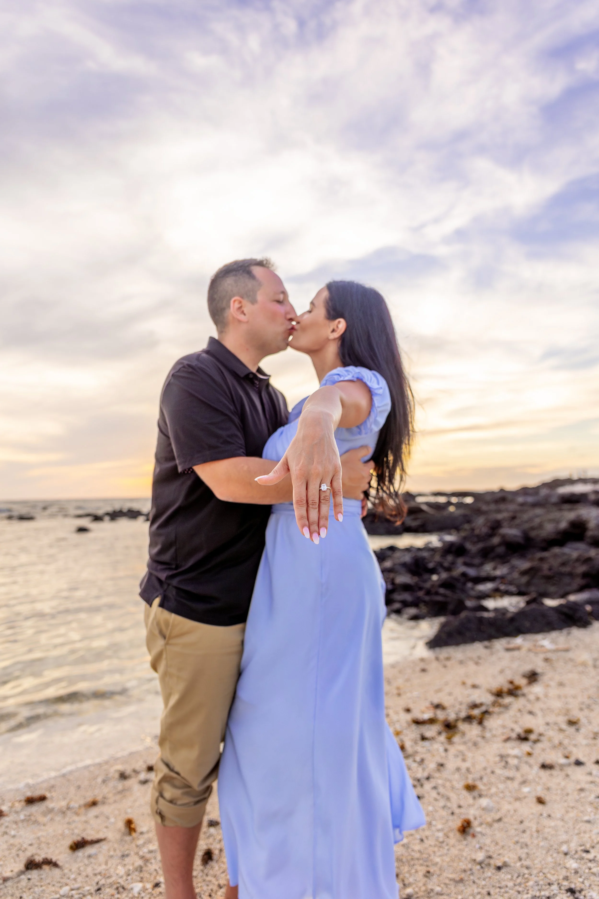 A couple sharing a kiss on the beach at sunset, with the woman showing her engagement ring.