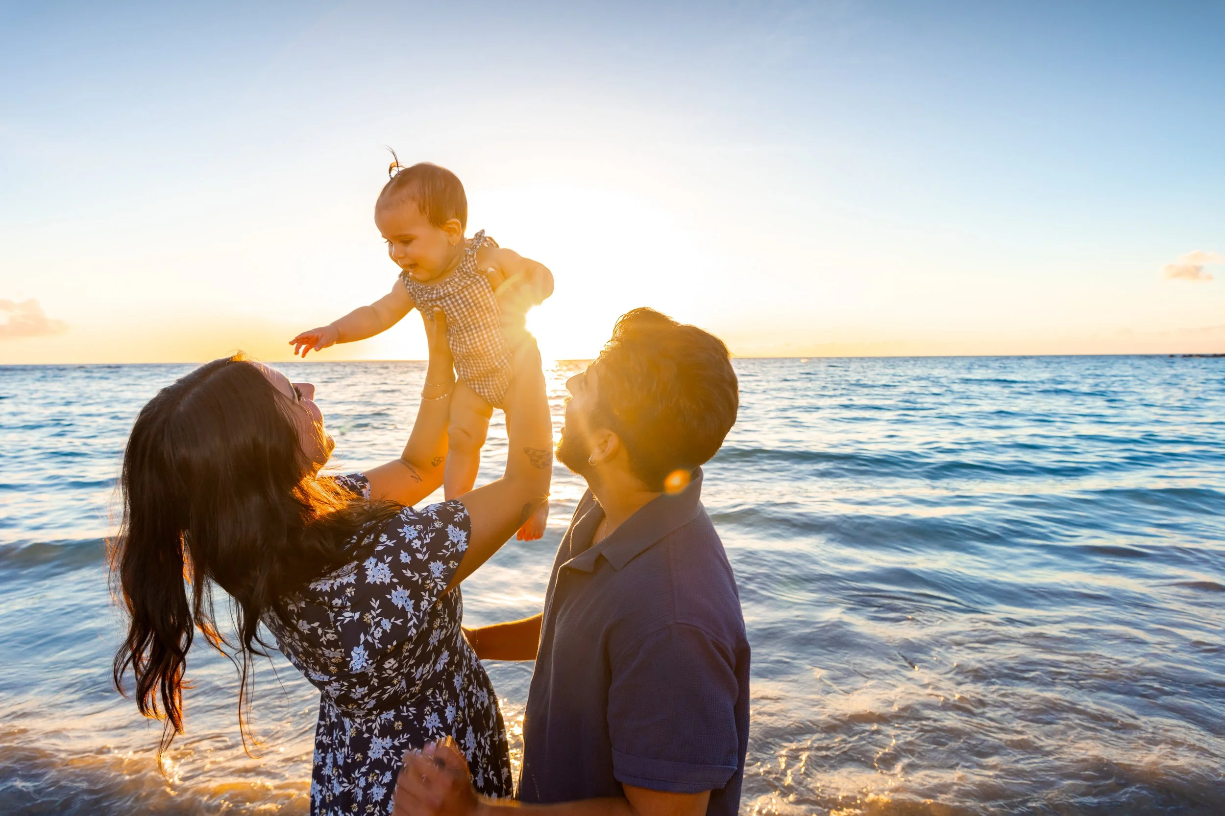 A family of three at the beach during sunset, with the mother lifting a young girl into the air while the father holds her legs, all smiling and enjoying the moment.