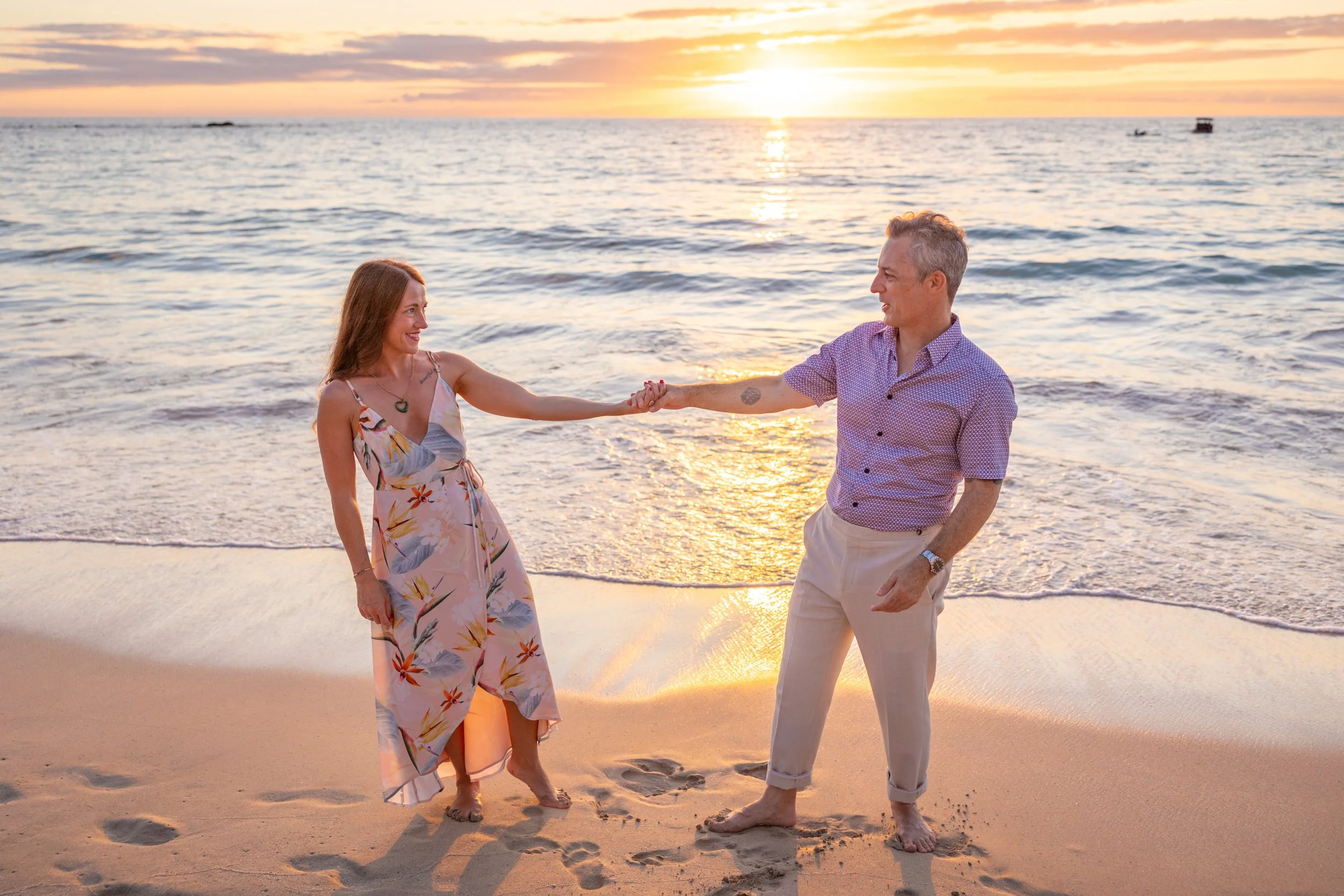 A couple holding hands on the beach during sunset, smiling at each other.
