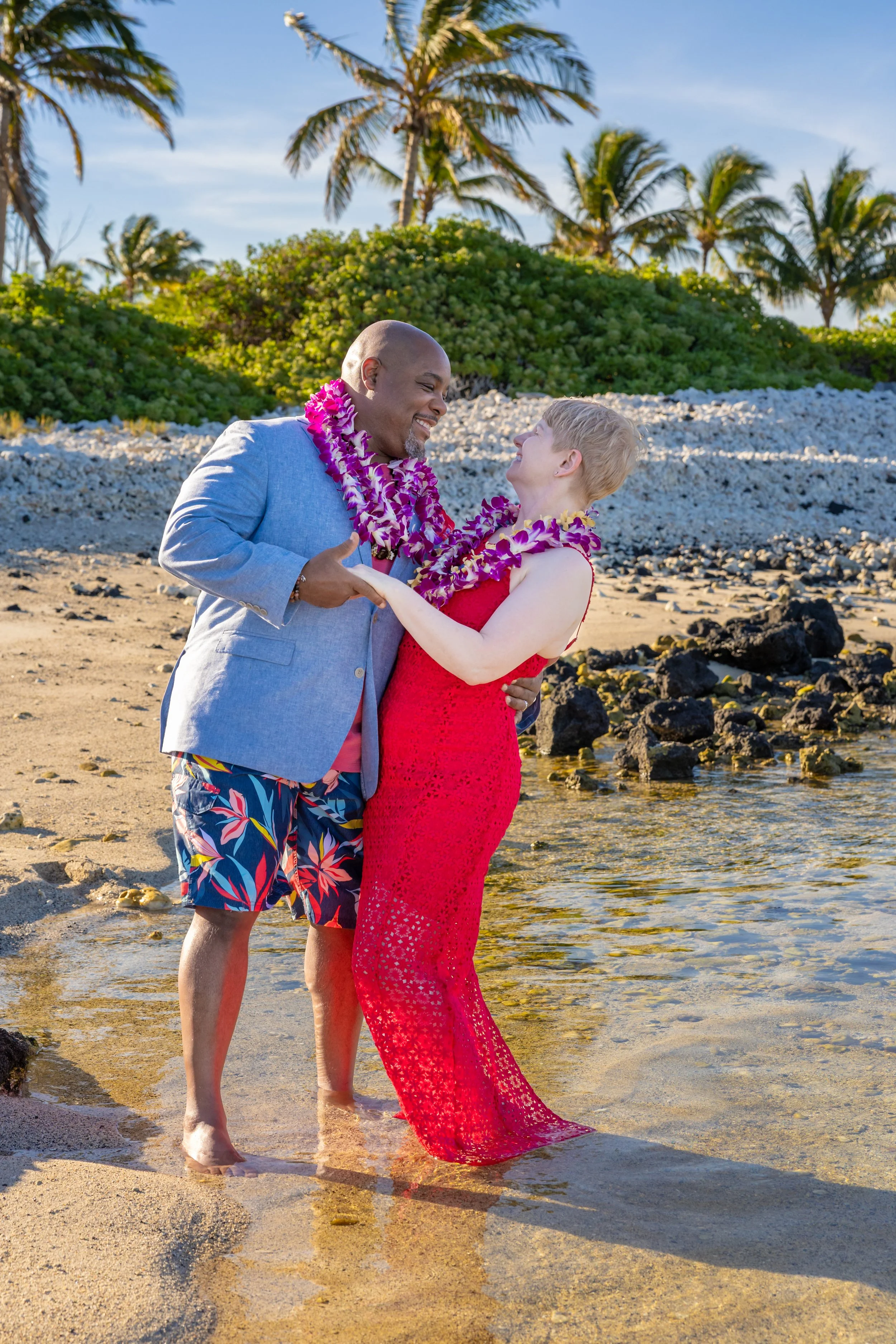 A couple dancing and smiling on a sandy beach with palm trees and clear blue sky in the background, wearing leis and colorful clothing.