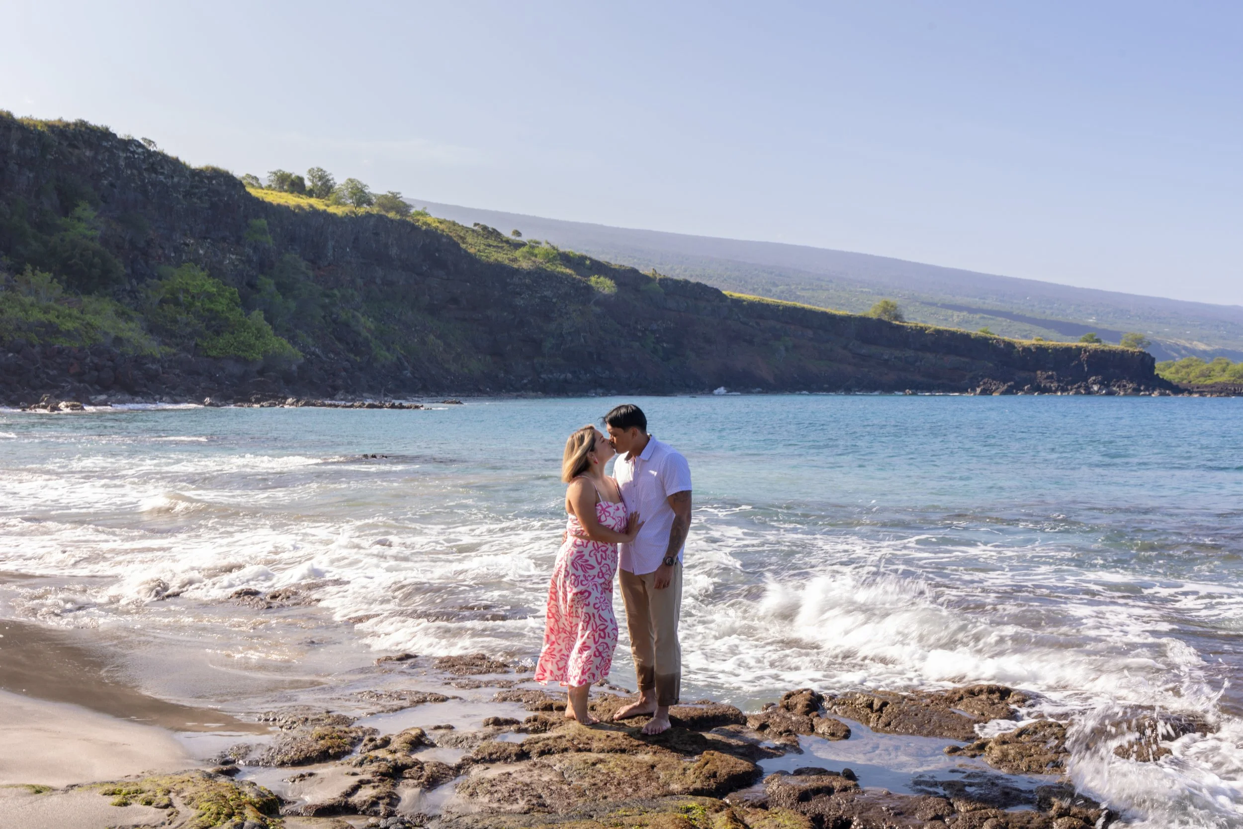 A couple standing on rocks at the beach, about to kiss, with waves and green hills in the background.
