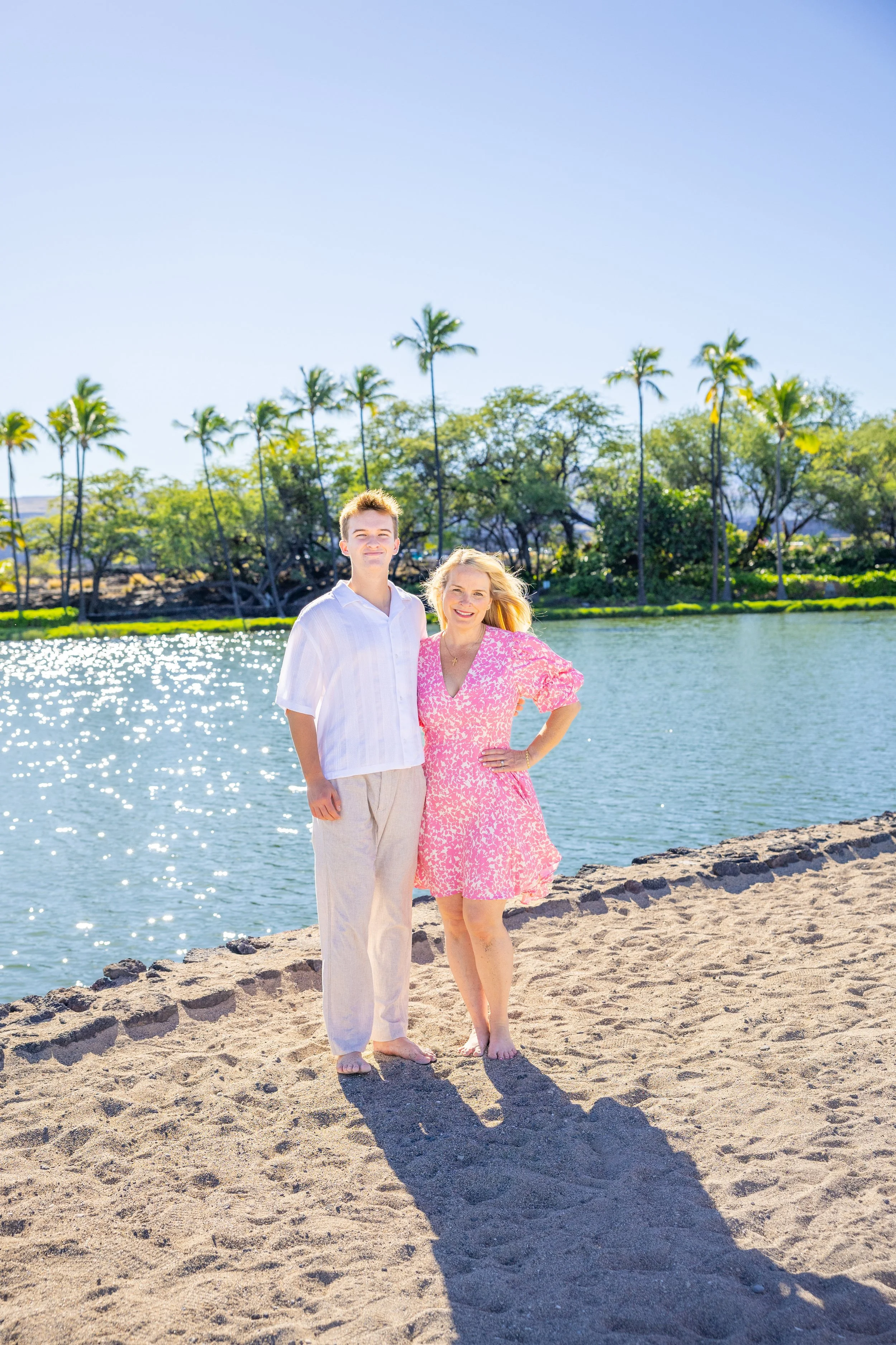 A woman and a young man standing on a sandy beach near a body of water, with palm trees and greenery in the background on a clear sunny day.
