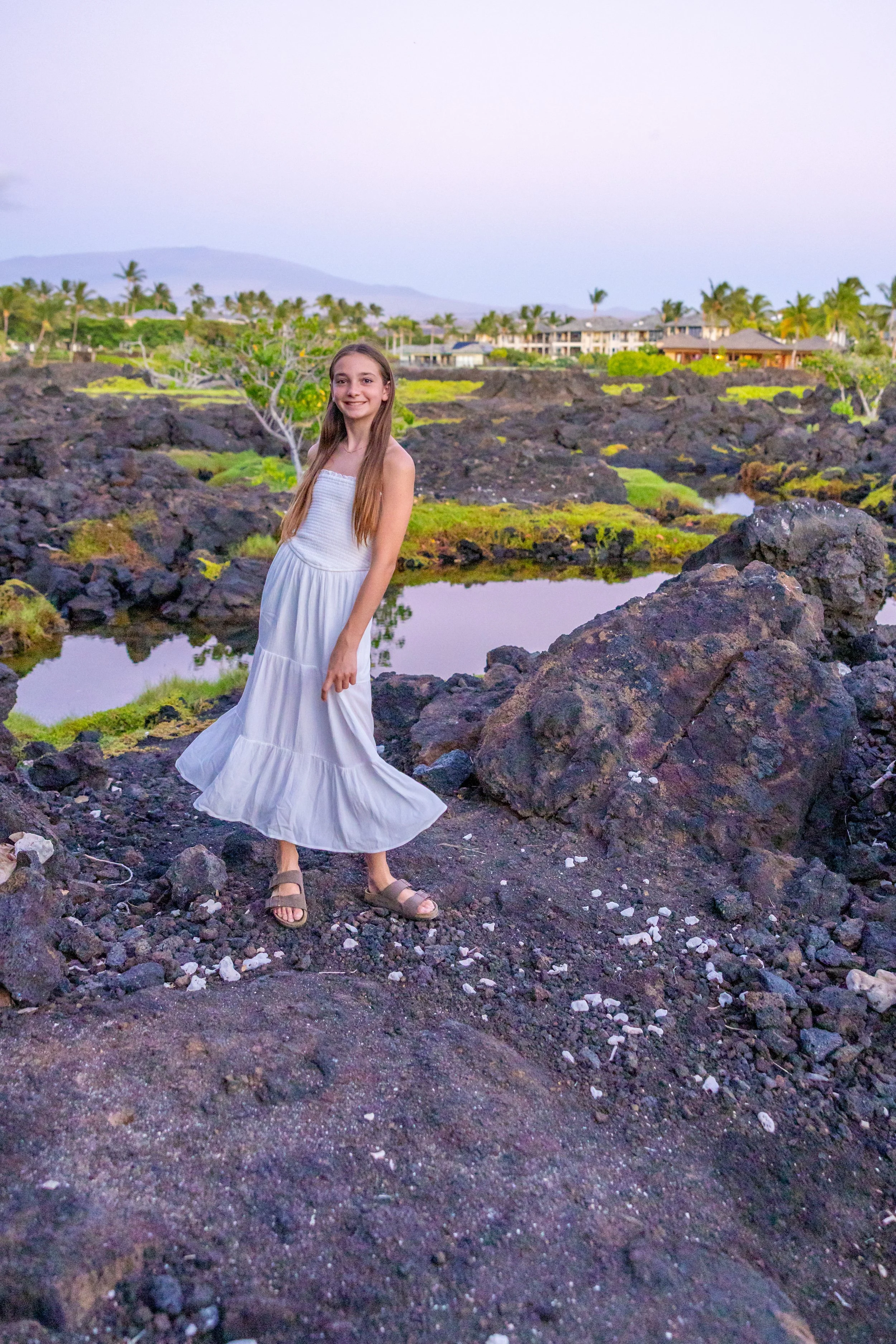 A girl in a white dress and sandals standing on volcanic rocks near small pools of water with palm trees and houses in the background.