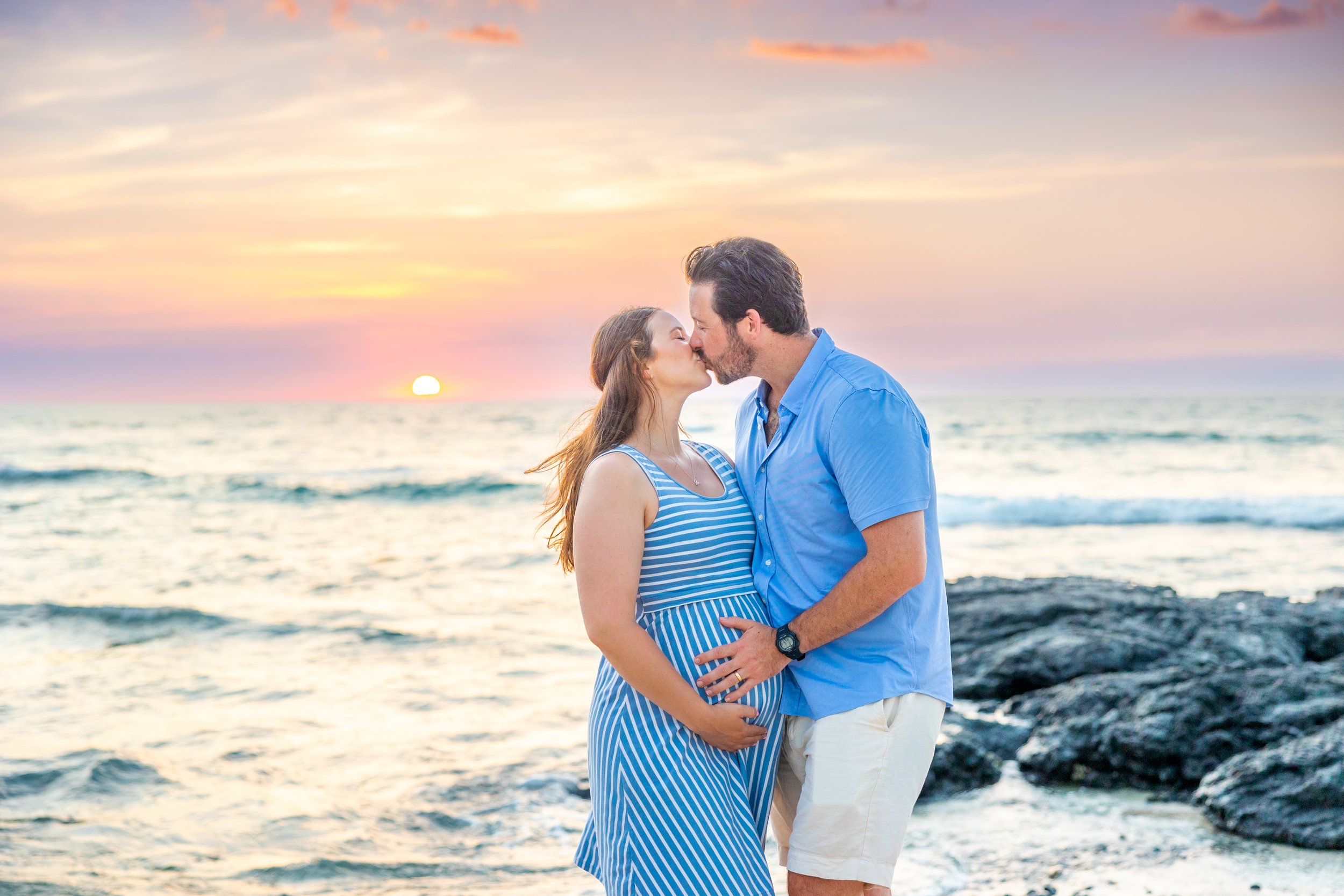 A pregnant woman and a man kiss on the beach during sunset.