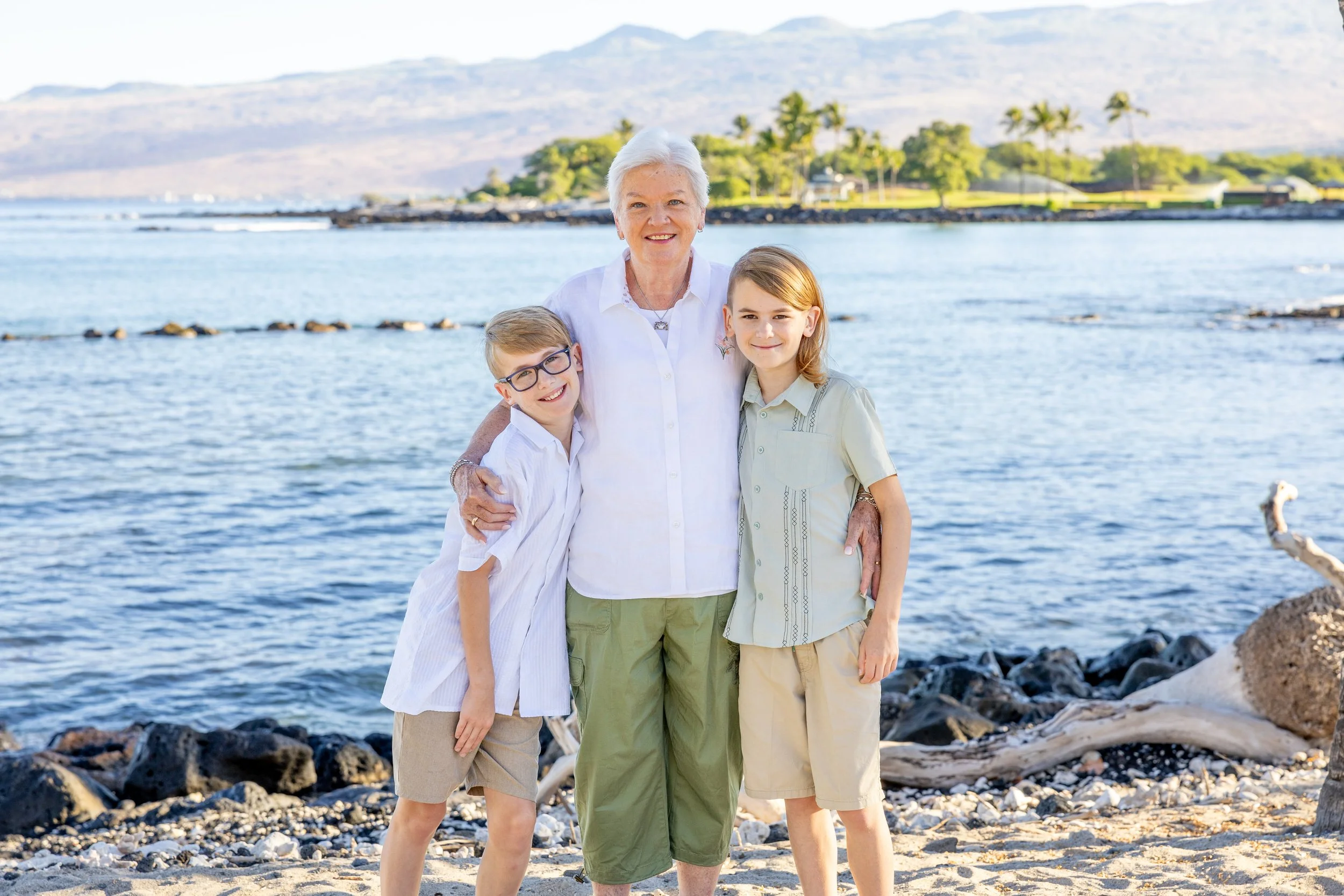 An elderly woman with two young children standing on a beach near the water, smiling and posing for a photo with mountains and palm trees in the background.