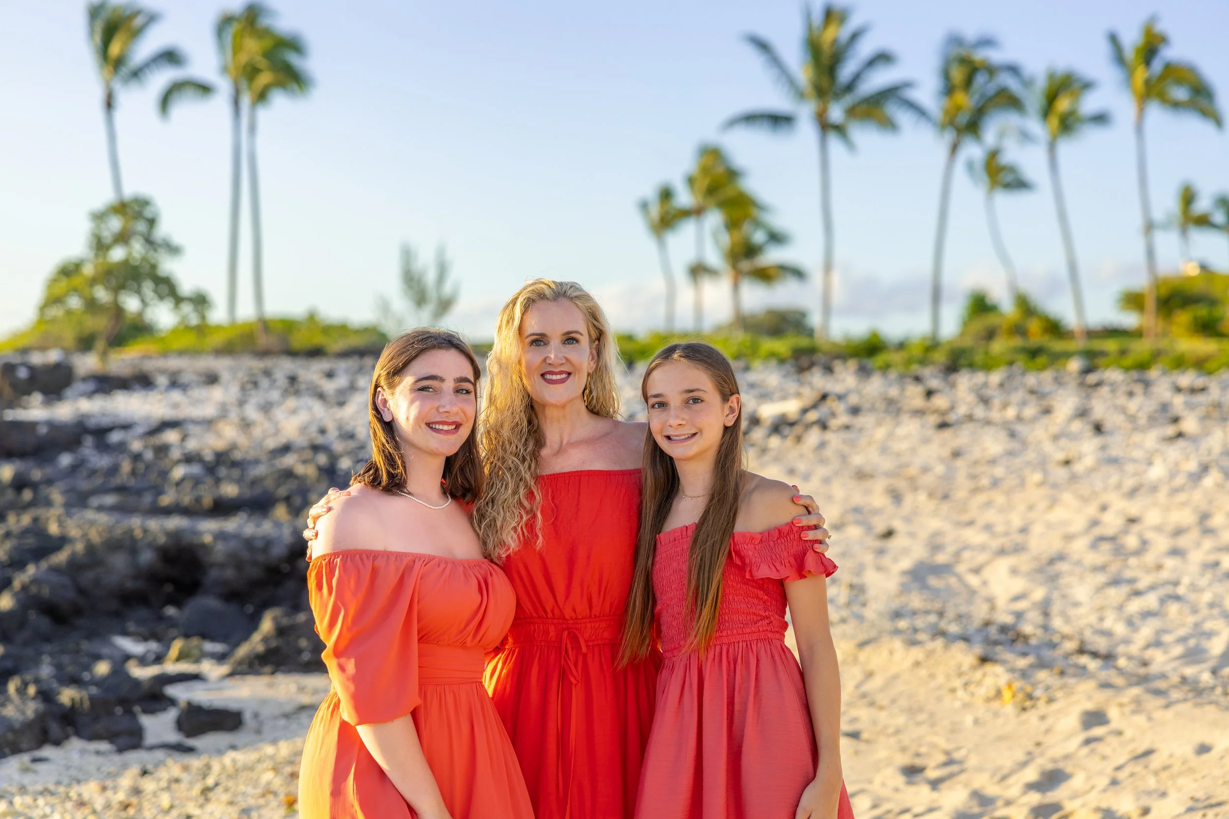 Three women in red dresses standing on a beach with palm trees in the background.